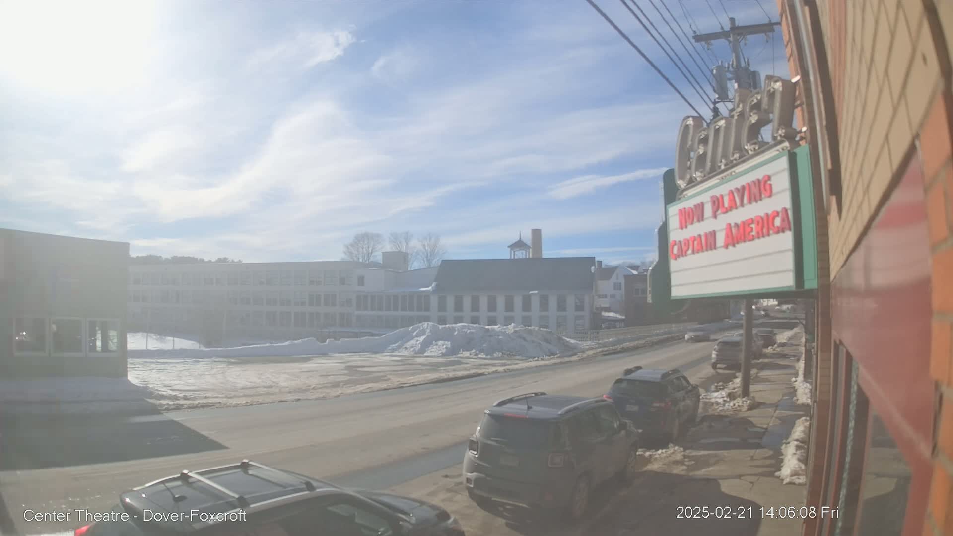 A sunny day shows a street scene with several cars parked and driving past snow piles and buildings, one of which is a movie theater marquee.