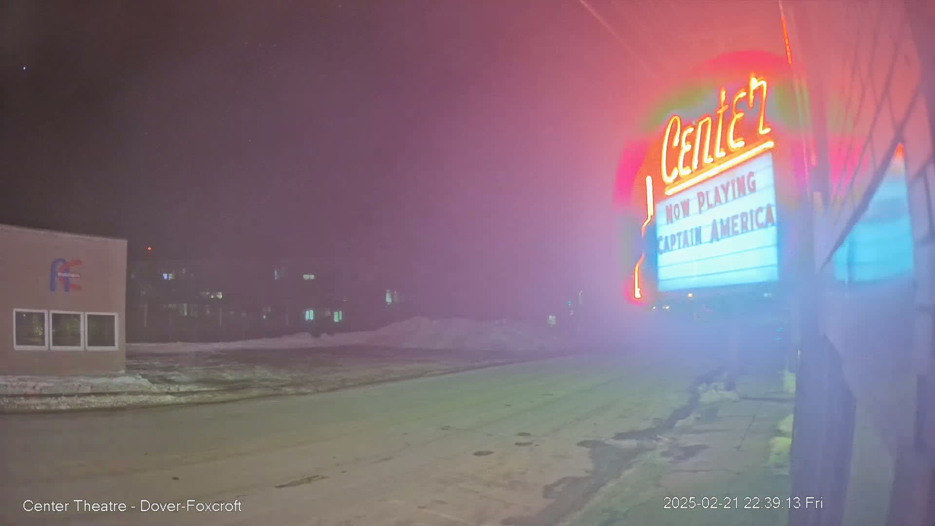 A fog-shrouded nighttime street scene shows a movie theater marquee advertising "Captain America," with a building and snow-covered ground visible in the distance.