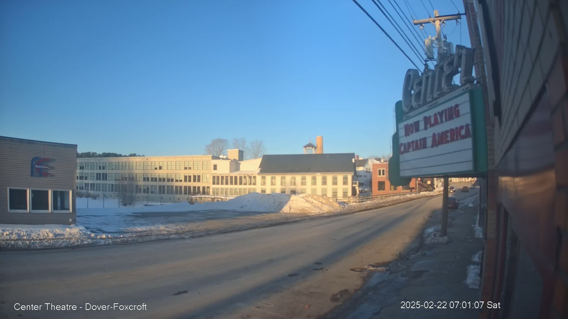 Under a clear, bright blue sky, a street scene shows several buildings, some with snow on the ground and a partially snow-covered parking lot.