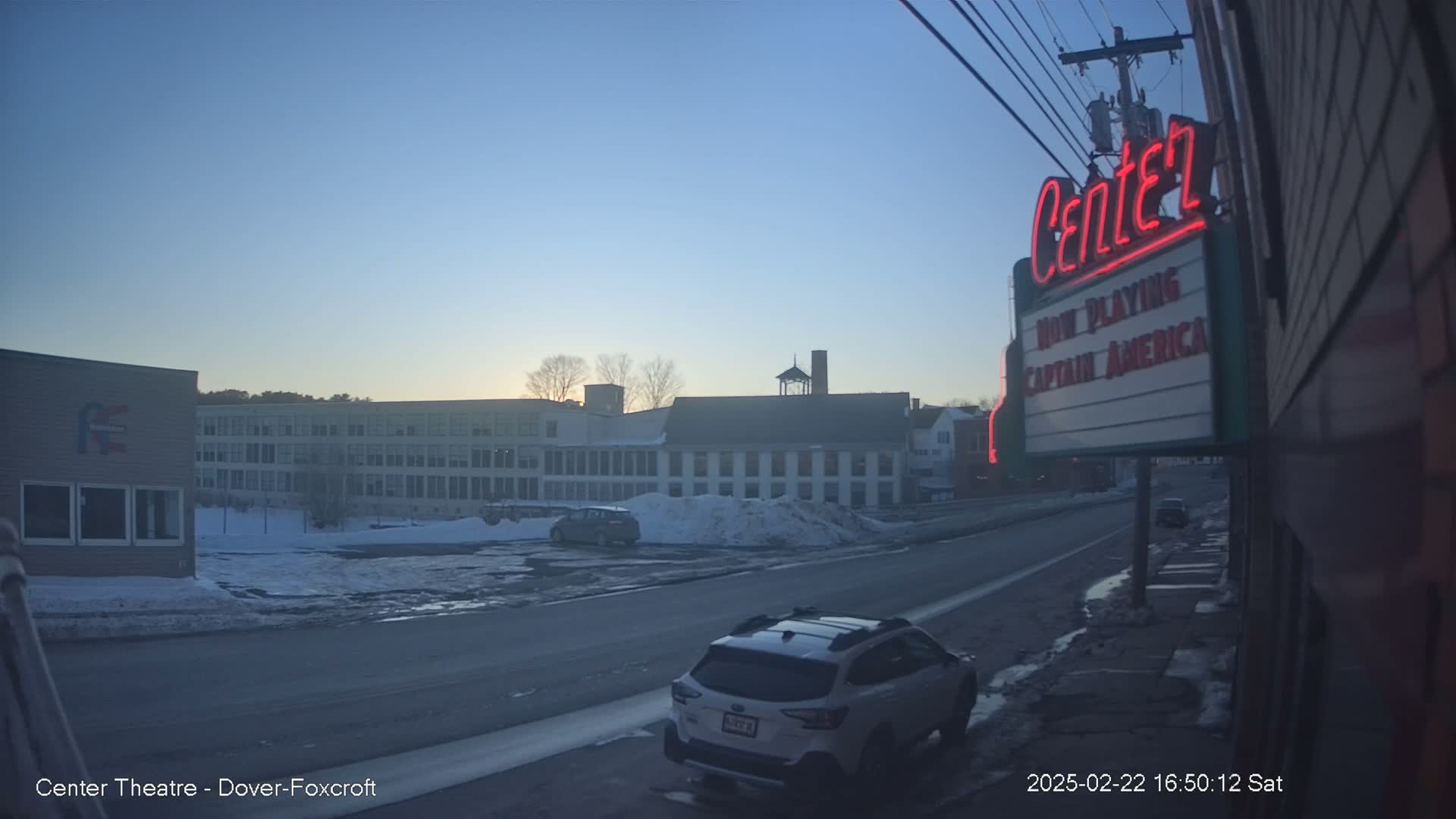 A street scene at sunset shows a snowy road with a white SUV parked near a movie theater marquee, and several buildings in the background.