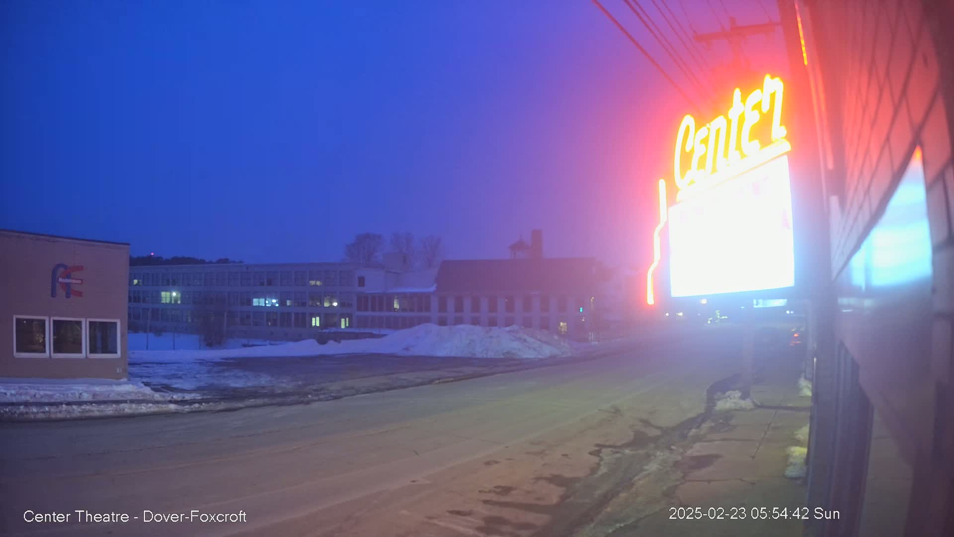 A brightly lit "Center" sign is visible near a snow-covered street at dusk, with buildings in the background under a hazy sky.