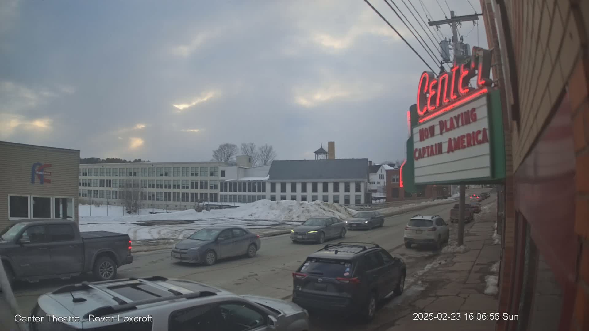 A street scene on an overcast day shows several cars driving on a snow-covered road past a large building and a movie theater marquee advertising "Captain America."