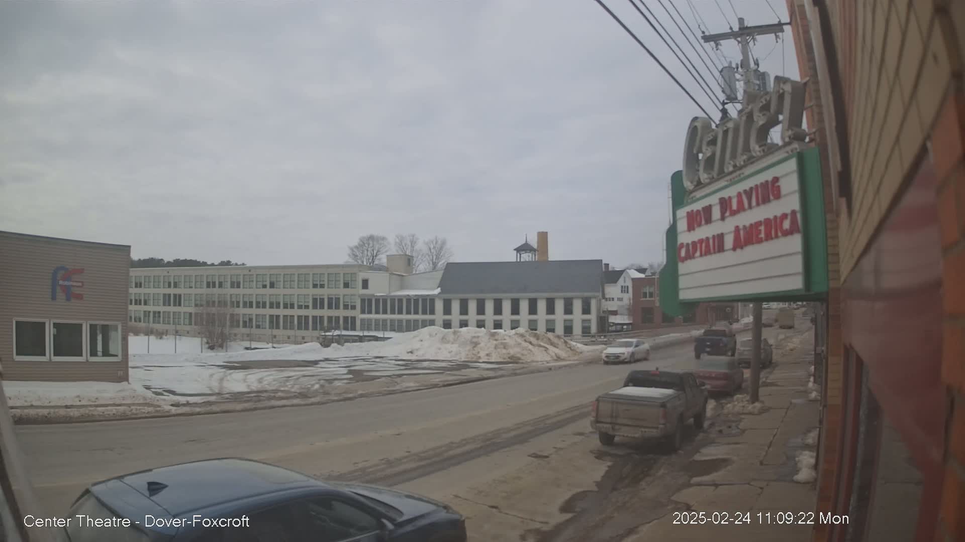 A street scene on an overcast day shows several vehicles parked and driving along a road in front of buildings, including a large multi-story structure and a smaller building with a marquee.  There is snow piled on the sides of the street.