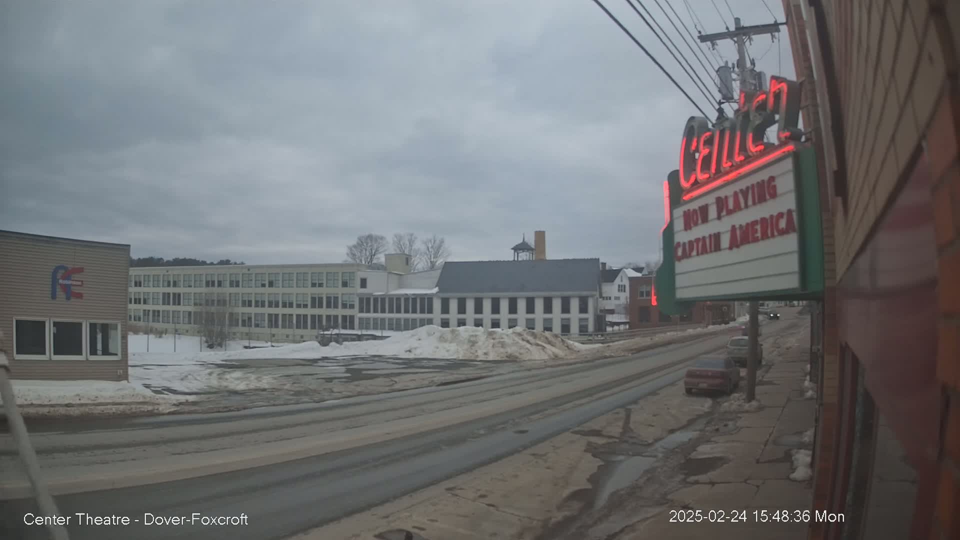 A street scene on an overcast day shows a multi-story building, smaller buildings, and a movie theater marquee advertising Captain America, with some snow and ice patches on the ground.