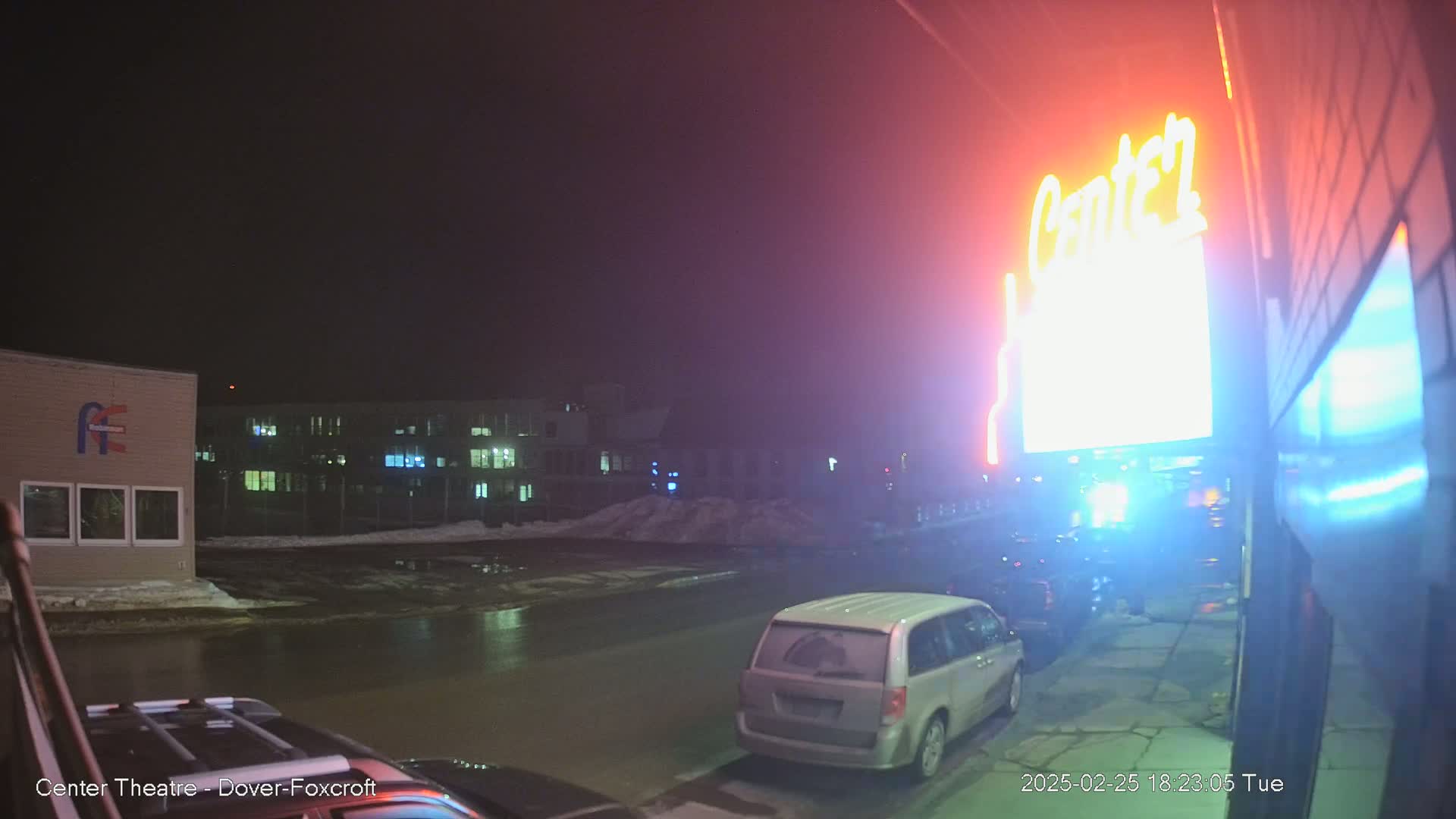 A nighttime street scene shows a minivan parked near a building with a brightly lit sign, under overcast skies with wet pavement.