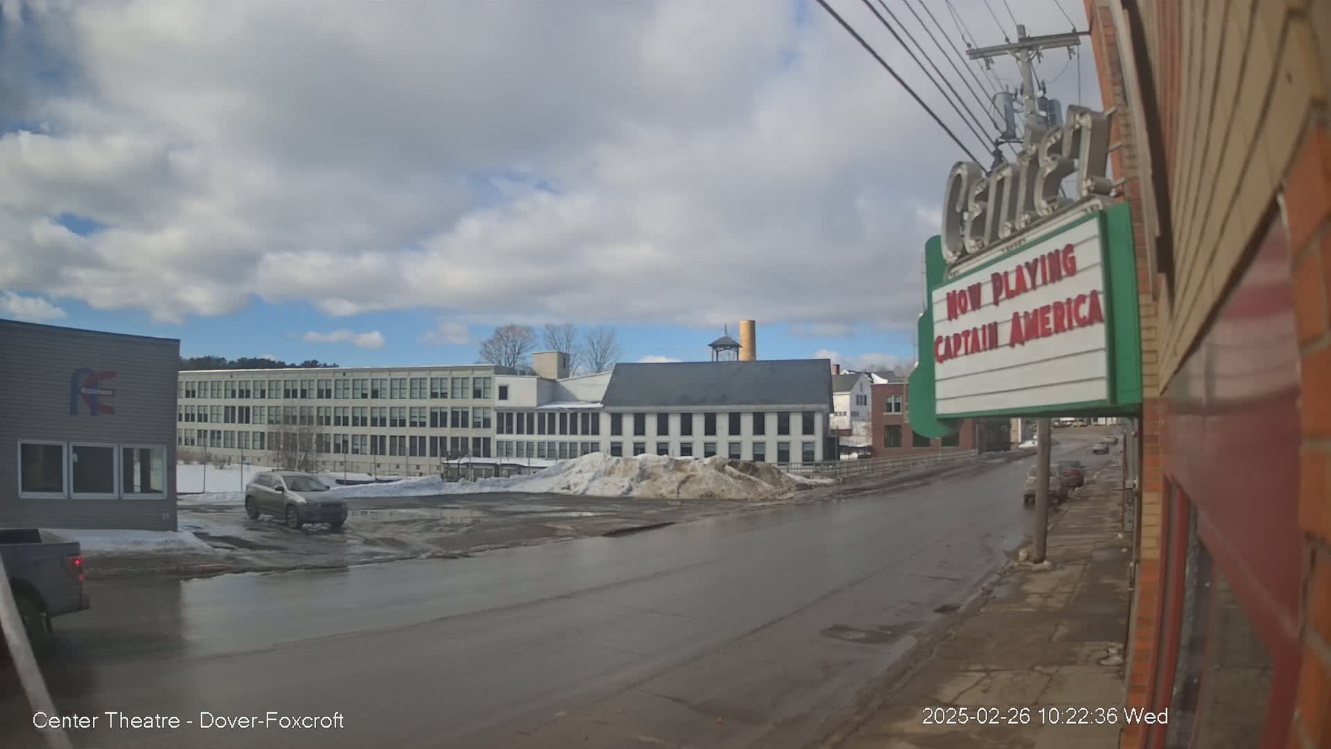 A partly cloudy day reveals a street scene with several buildings, including a large factory and a movie theater marquee advertising Captain America, some cars, and patches of snow and ice on the ground.