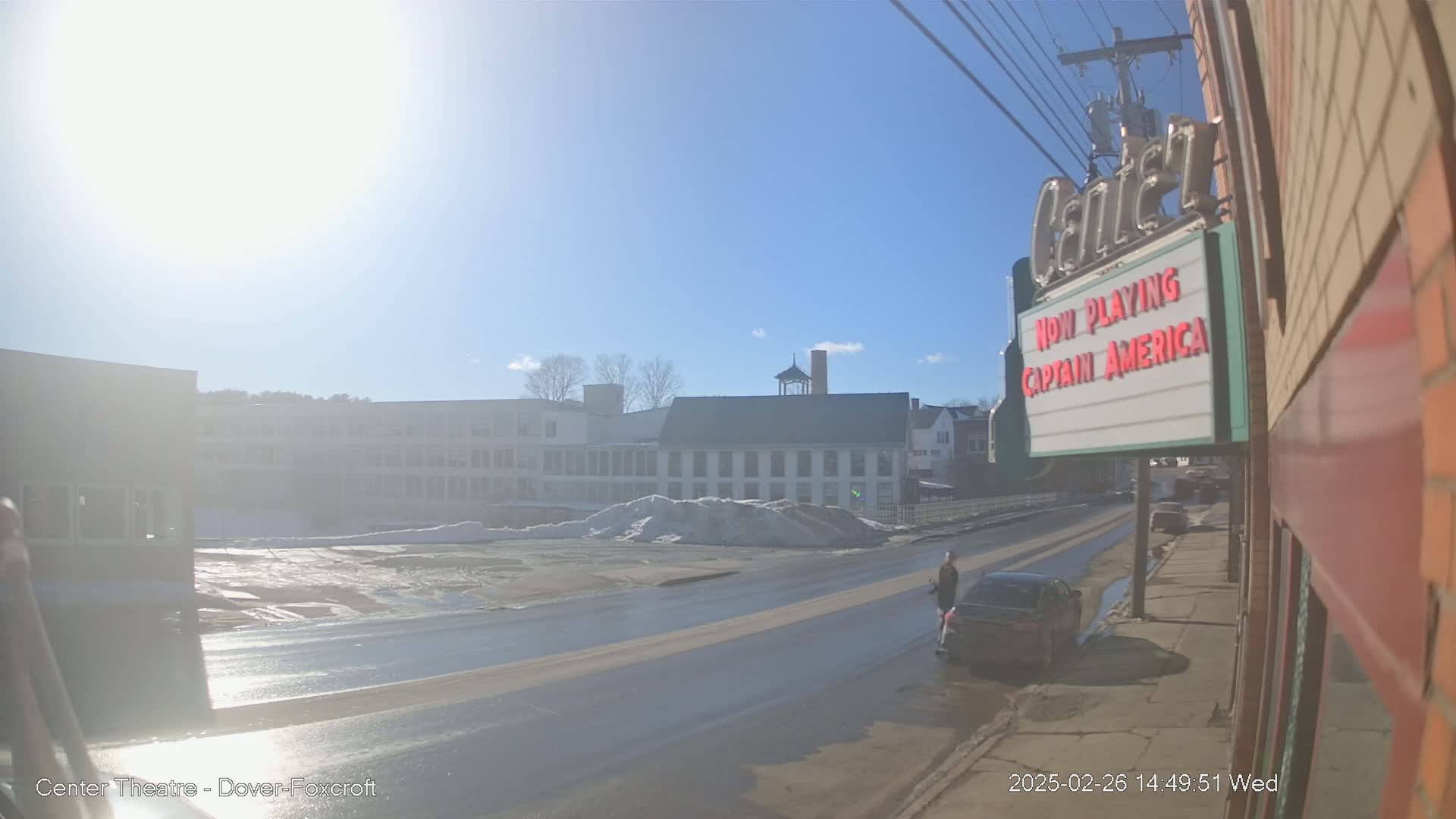 A sunny day reveals a street scene with a few cars, a pedestrian, and a snow pile in front of several buildings, one of which is a theater.