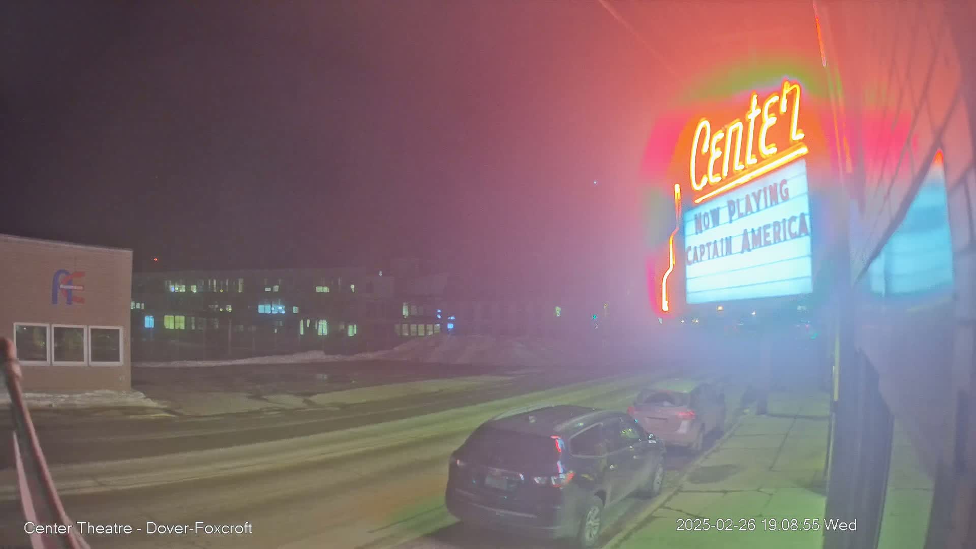 A foggy night scene shows a movie theater marquee advertising Captain America, with cars parked along a snow-dusted street in front of it.