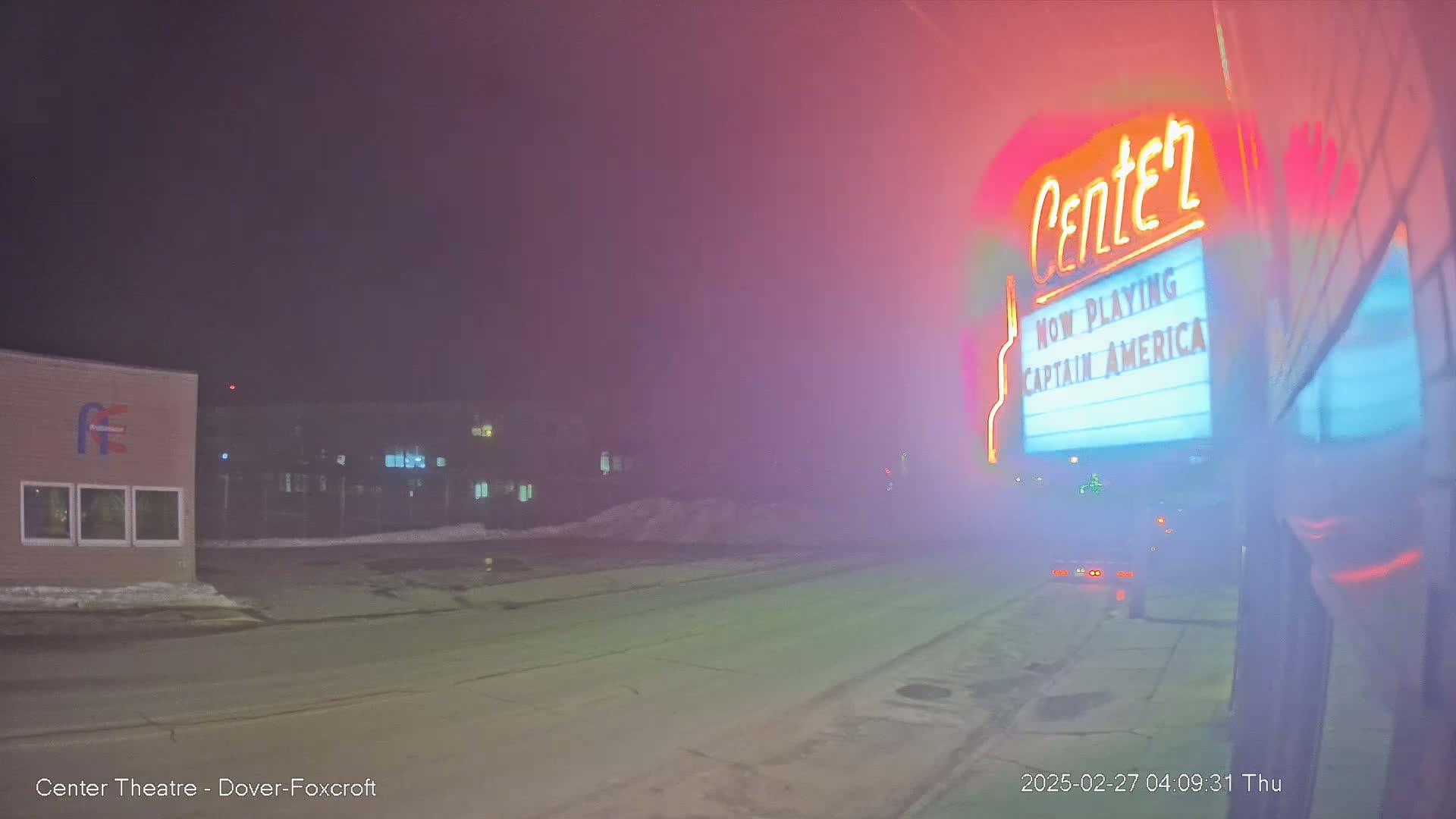 A fog-shrouded street at night features a movie theater marquee advertising "Captain America," with buildings and snow visible in the background.