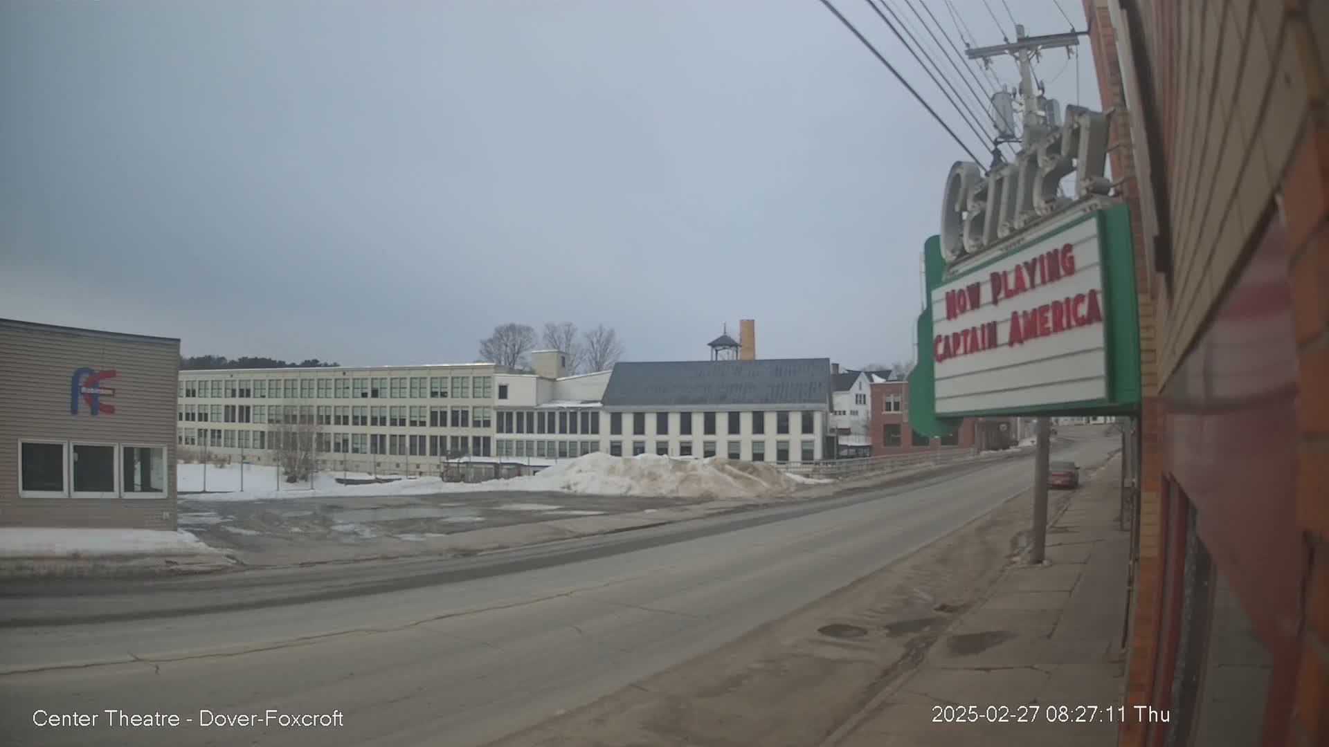 A street scene on an overcast day shows a road leading past industrial buildings and a movie theater marquee advertising Captain America.