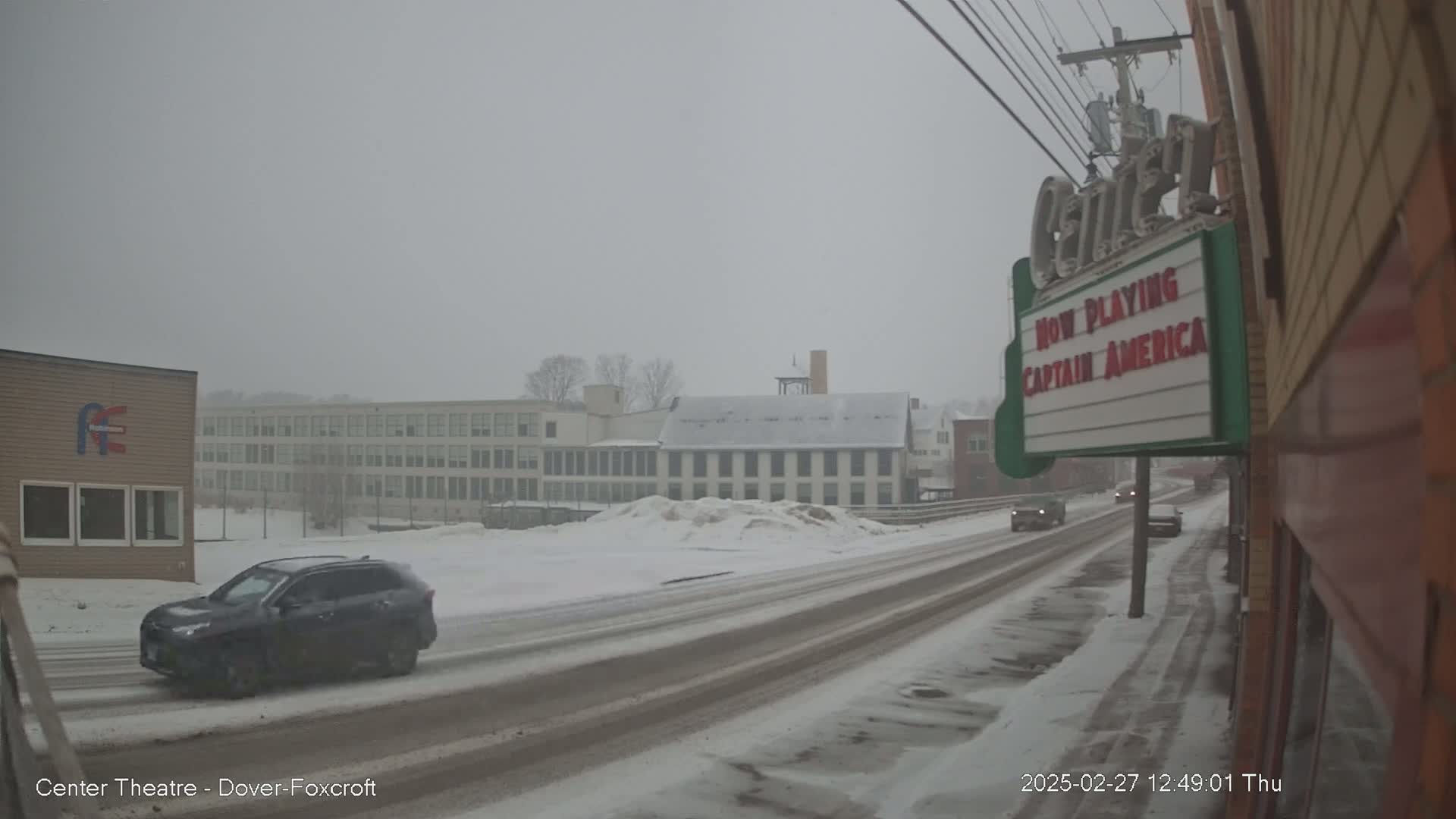 A snow-covered street scene shows a dark-colored SUV driving past buildings, including a multi-story structure and a movie theater marquee advertising Captain America.