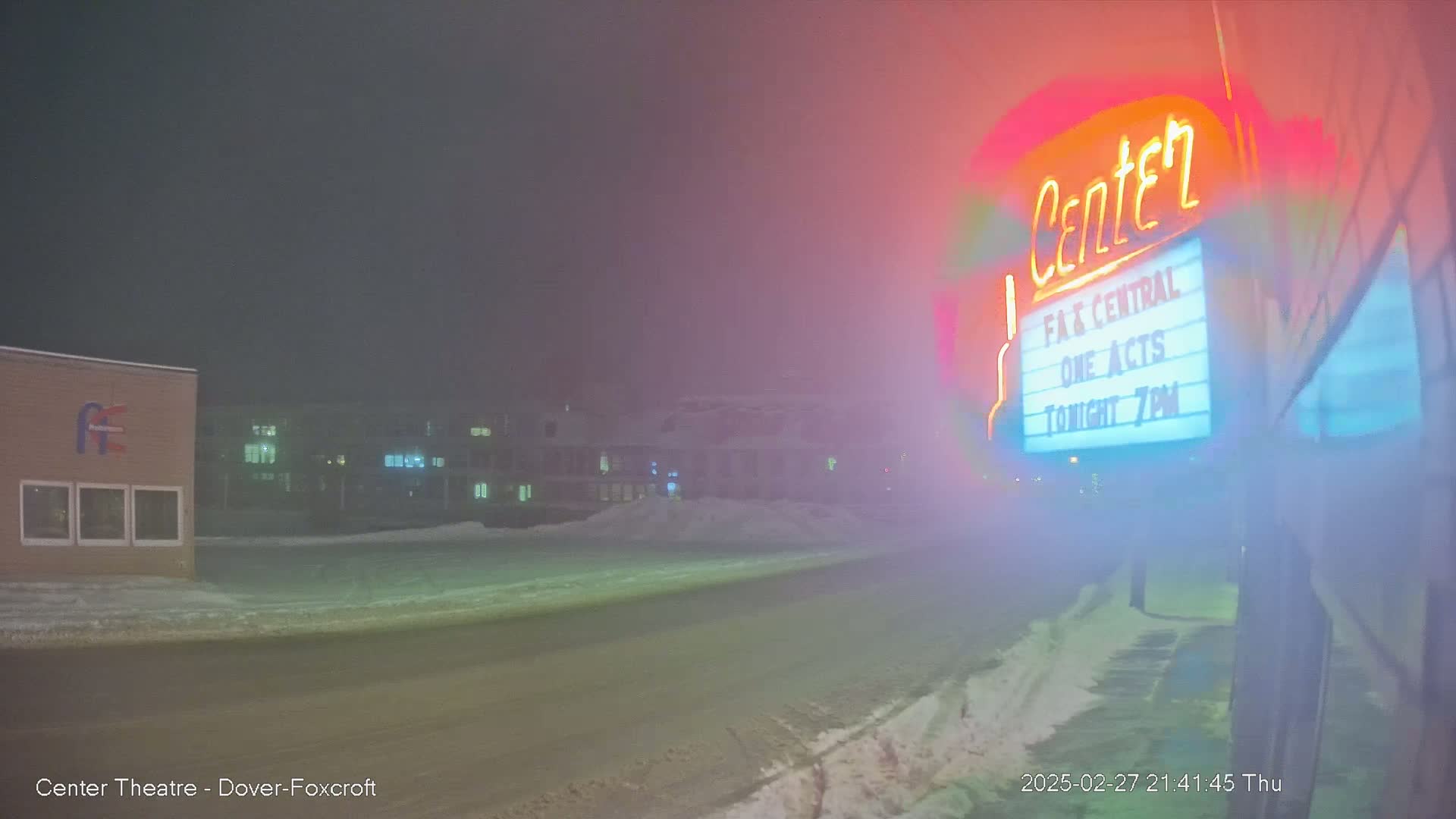 A foggy night scene shows a snow-covered street in front of a theater with a neon sign.