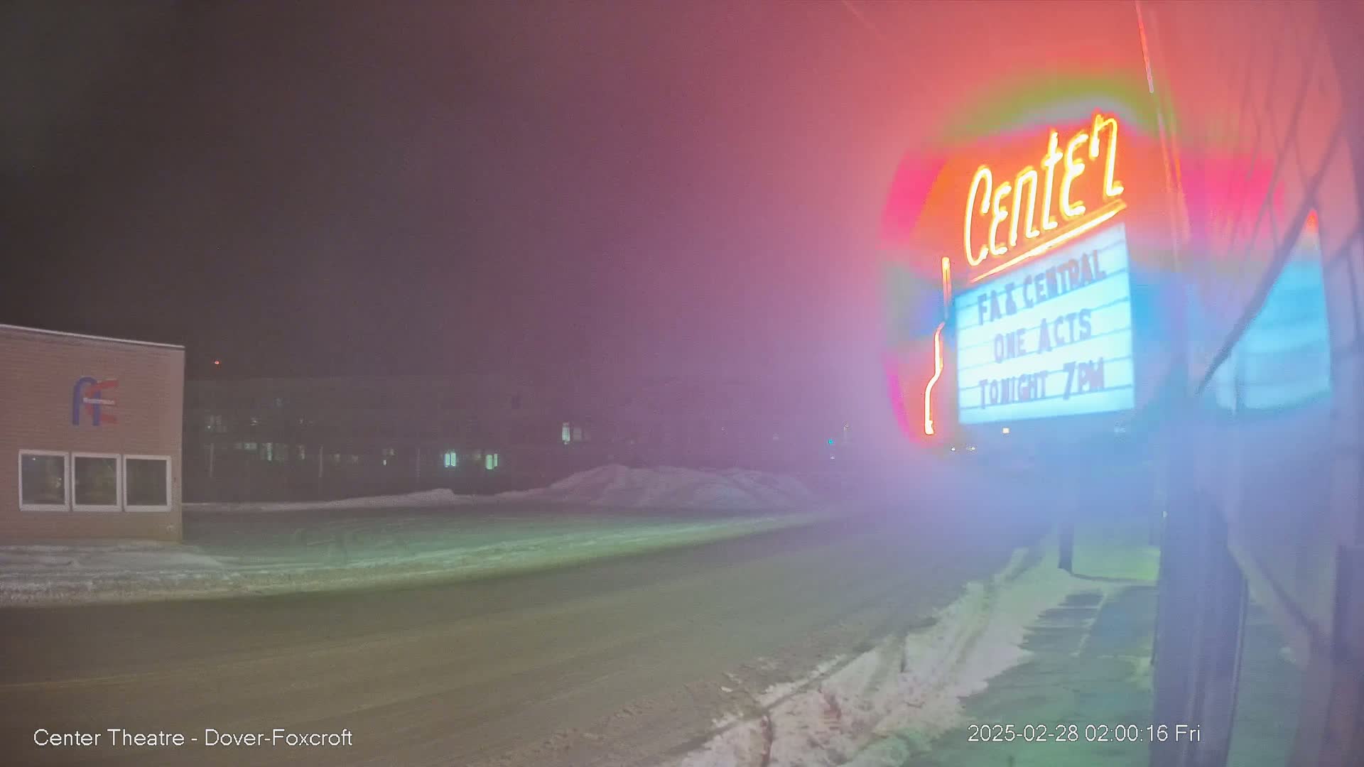 A brightly lit theater marquee in a foggy, snowy night scene shows a road and buildings in the background.