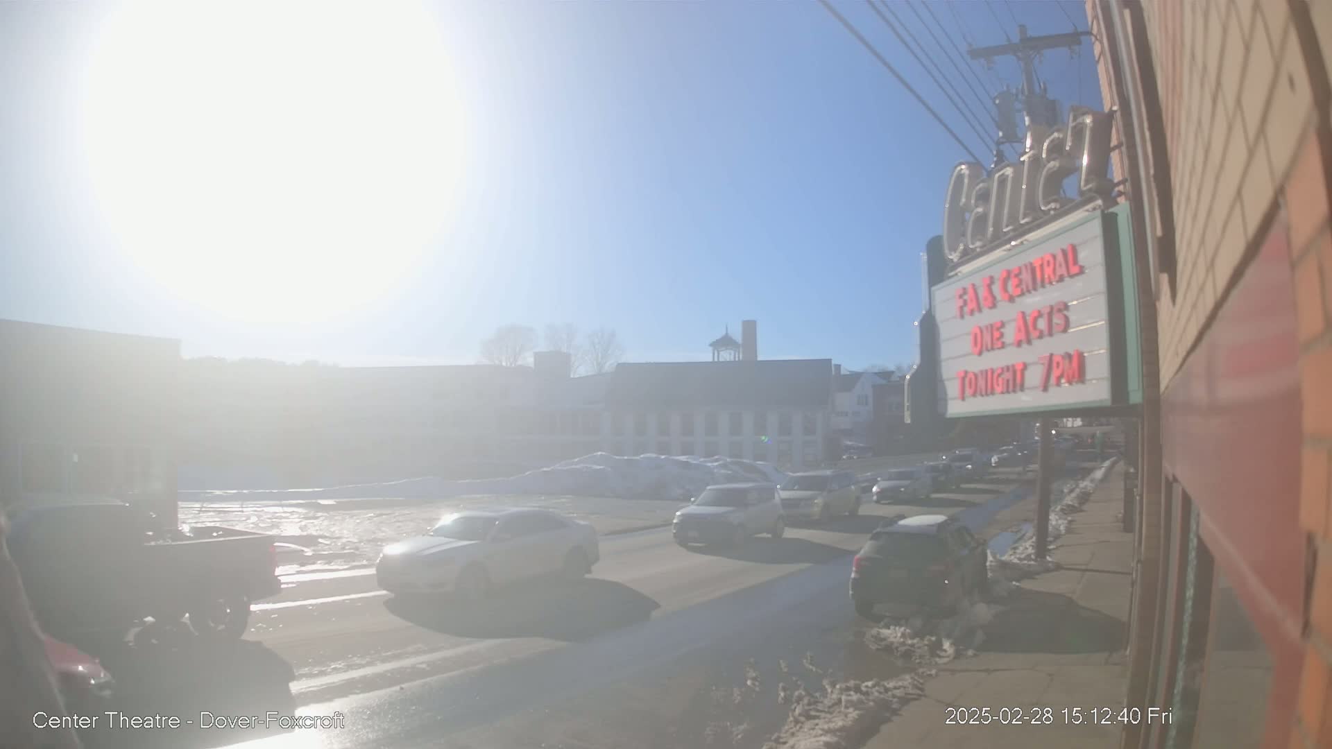 A bright, sunny day shows a street scene with several cars driving past a theater marquee and snow banks lining the road.