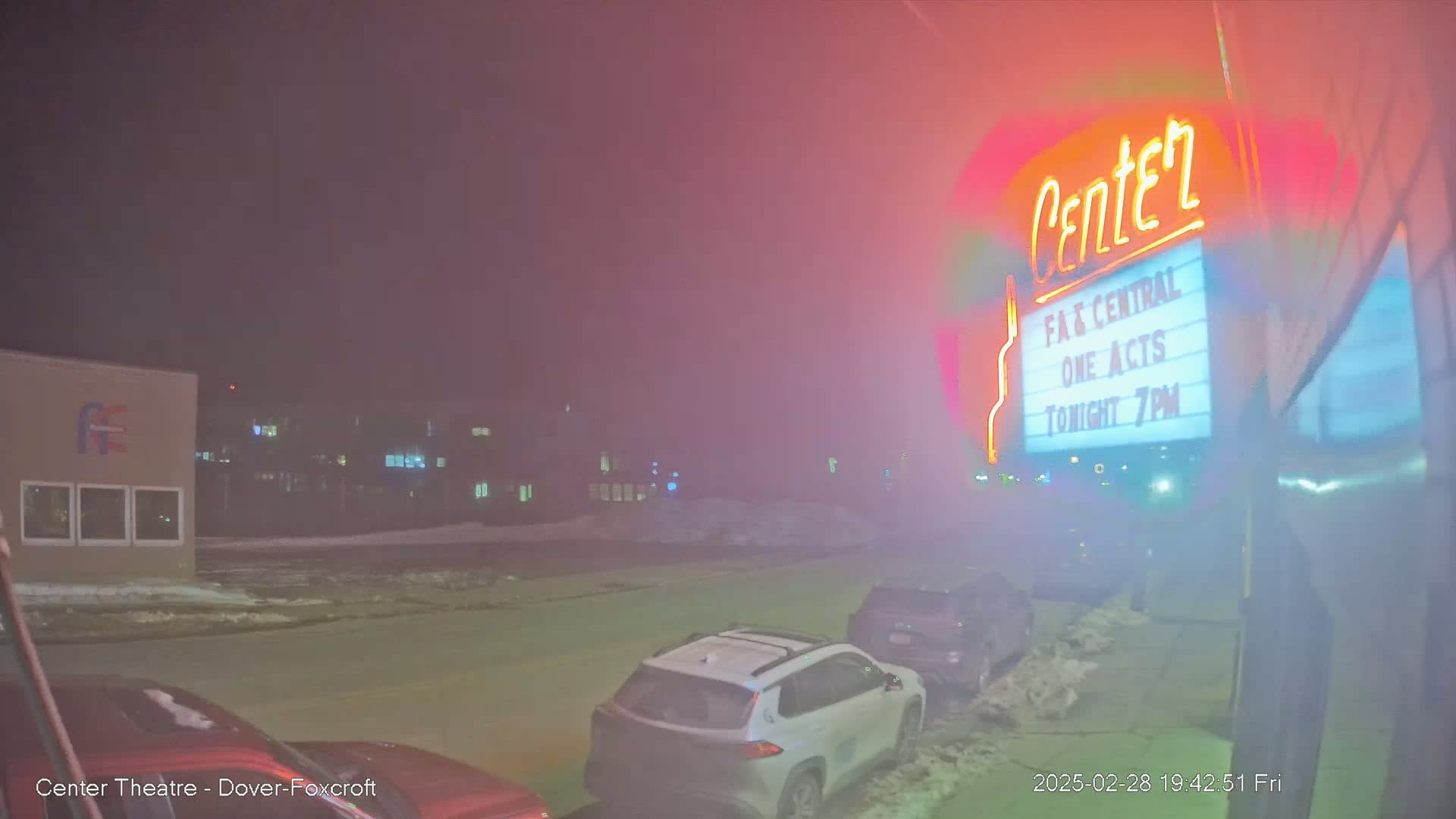A foggy night scene shows a theater marquee advertising a show, with cars parked along a street in front of buildings.