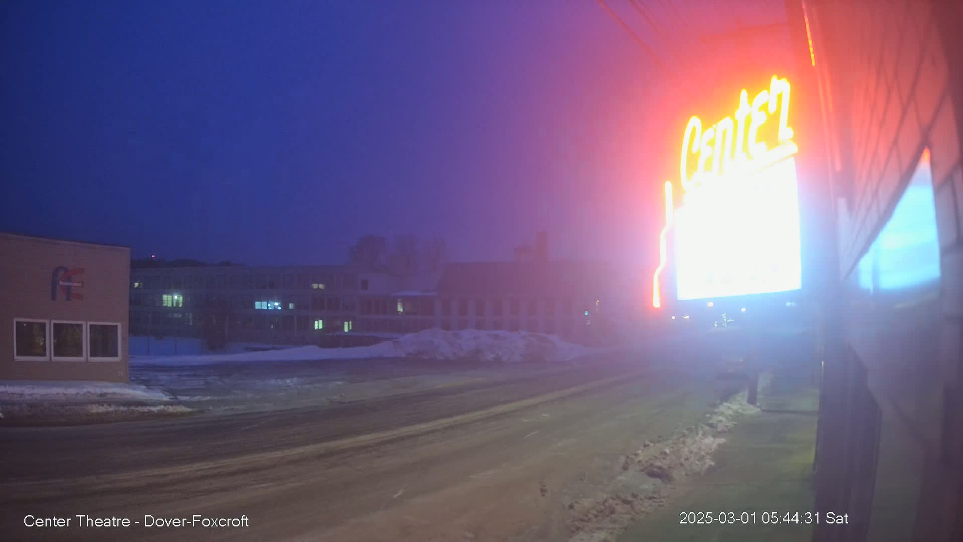 A foggy, dimly lit street scene at night shows a road, buildings covered in snow, and a neon sign.