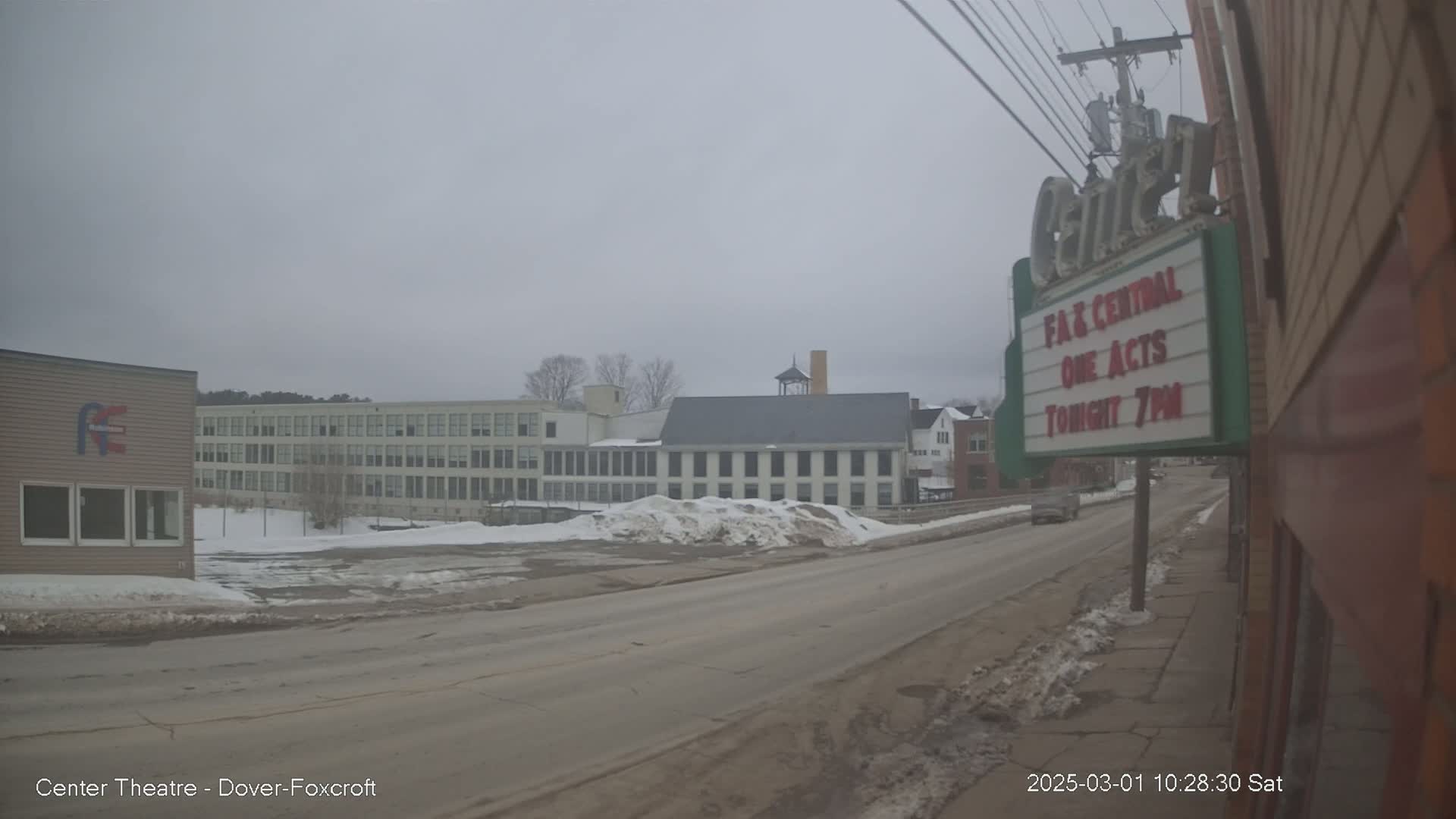 A mostly overcast day reveals a street scene with several buildings, including a large factory-like structure, with snow piled on the sides of the road.