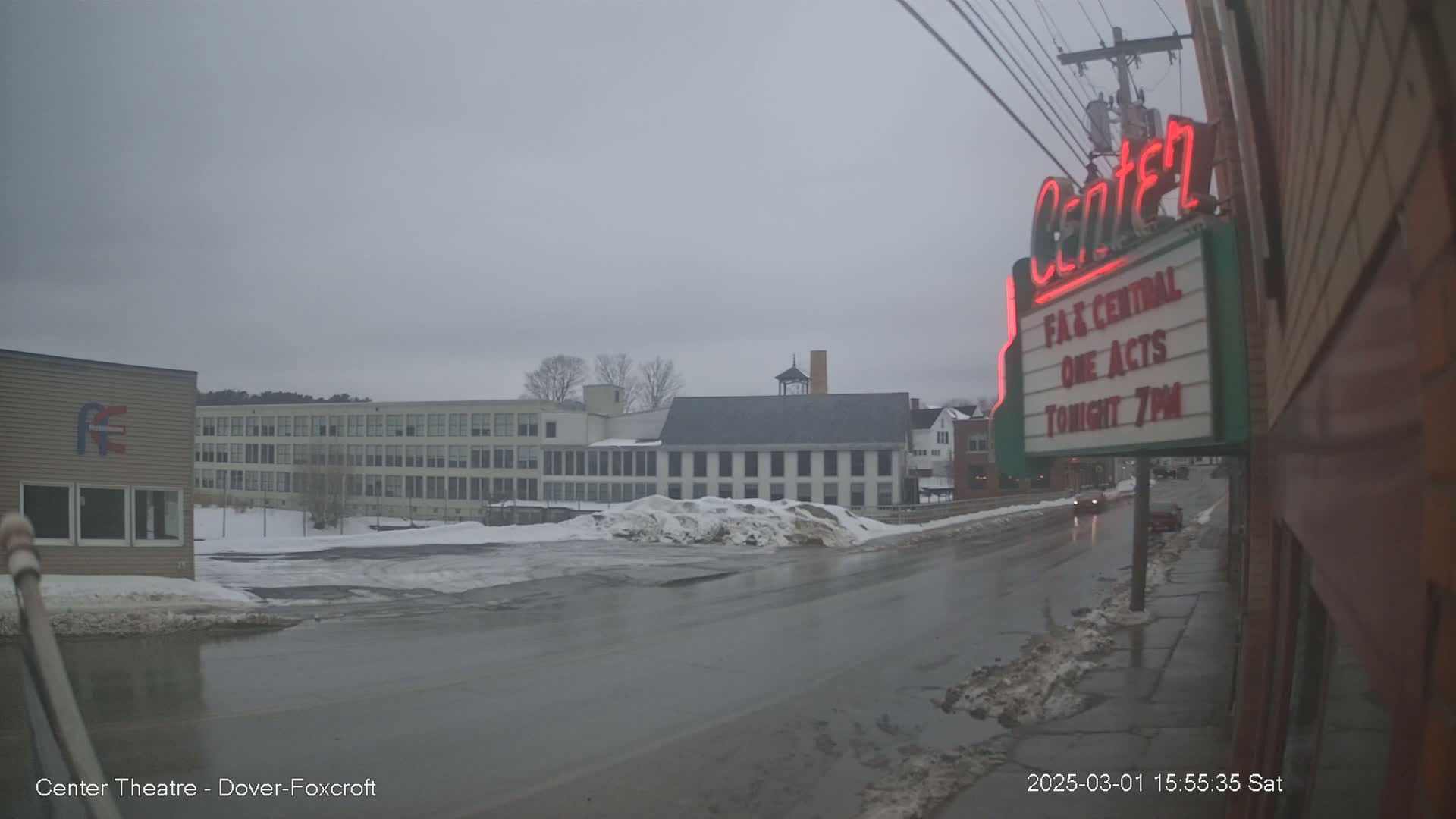 A wet street scene on an overcast day shows a theater marquee, a large building, and snow banks lining the road.