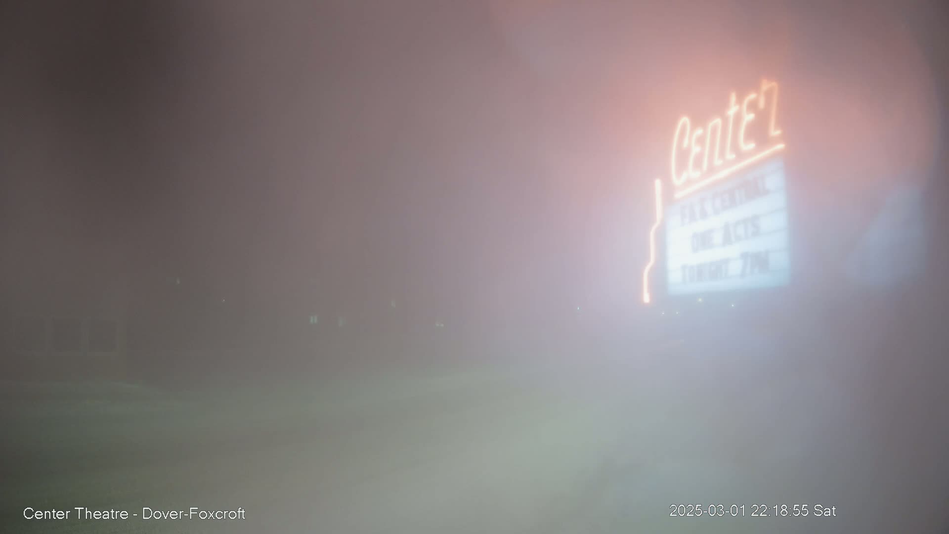 A dimly lit, fog-shrouded outdoor scene features a partially visible theater marquee with its name illuminated in neon.