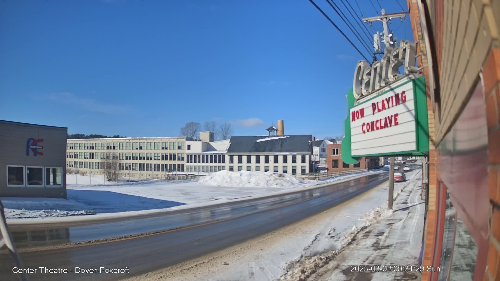 A sunny winter day shows a snow-covered street with several buildings, including a large factory and a theater marquee.