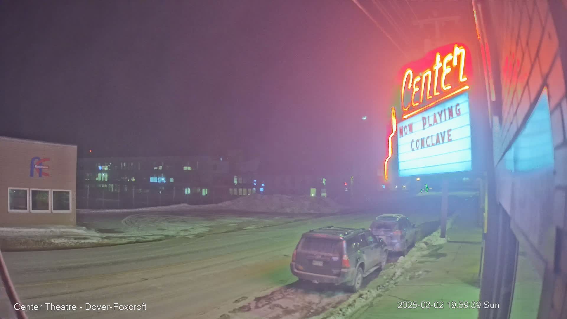 A hazy, snowy night scene shows a movie theater marquee with two cars parked in front, and other buildings visible in the background.