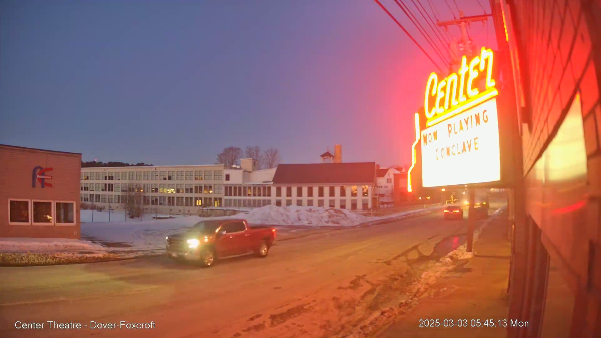 A red pickup truck drives past a movie theater marquee under a twilight sky with snow on the ground.