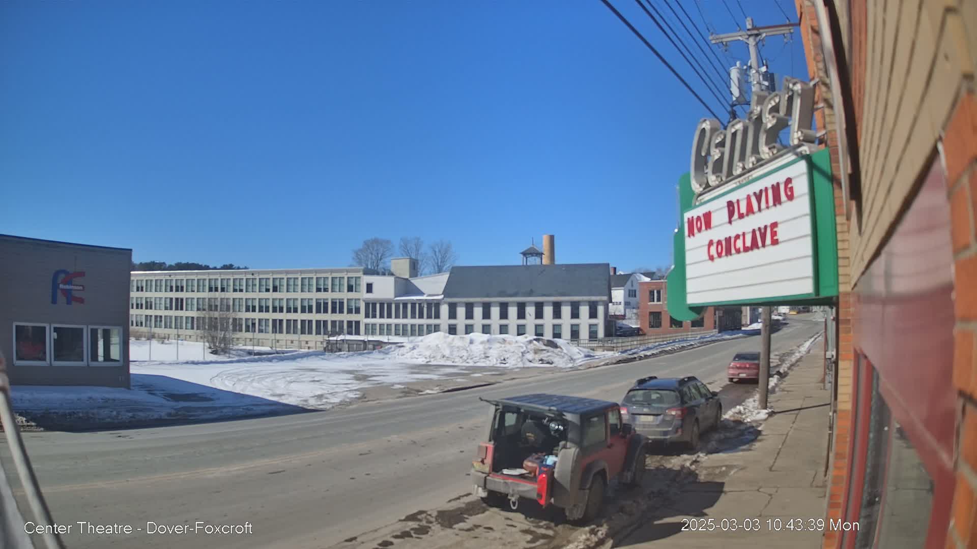 A sunny day reveals a street scene with several cars parked along a road, a large industrial building across the street, and a movie theater marquee advertising "Conclave."