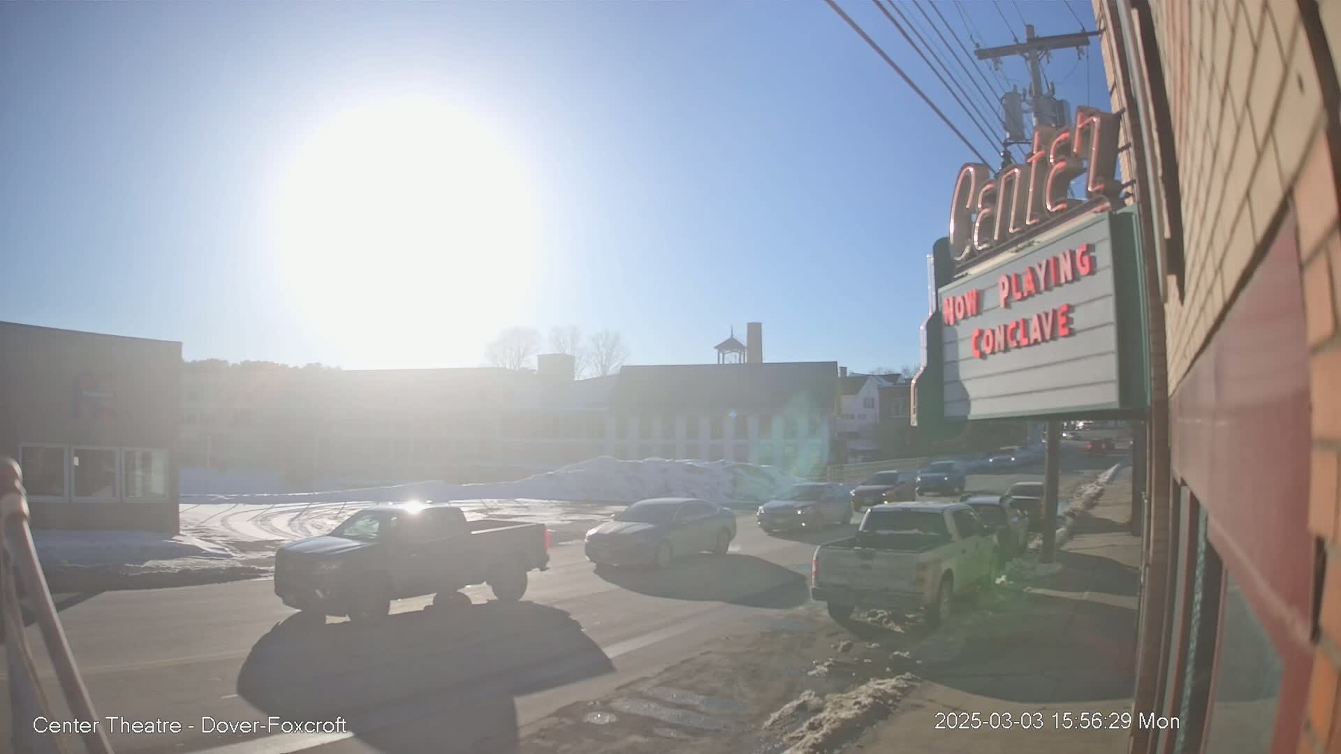 A sunny day shows several vehicles driving on a road in front of buildings, one of which is a movie theater with a marquee.