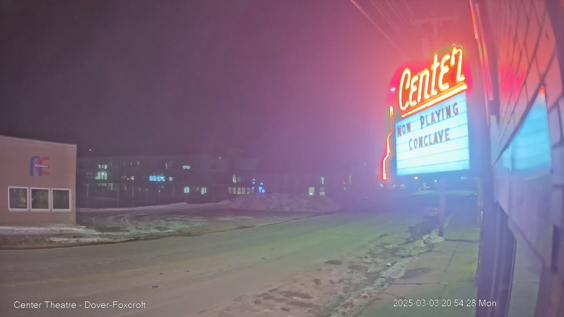 A nighttime, snowy scene shows a movie theater marquee with a film title and a building across the street under a hazy, pink-tinged sky.