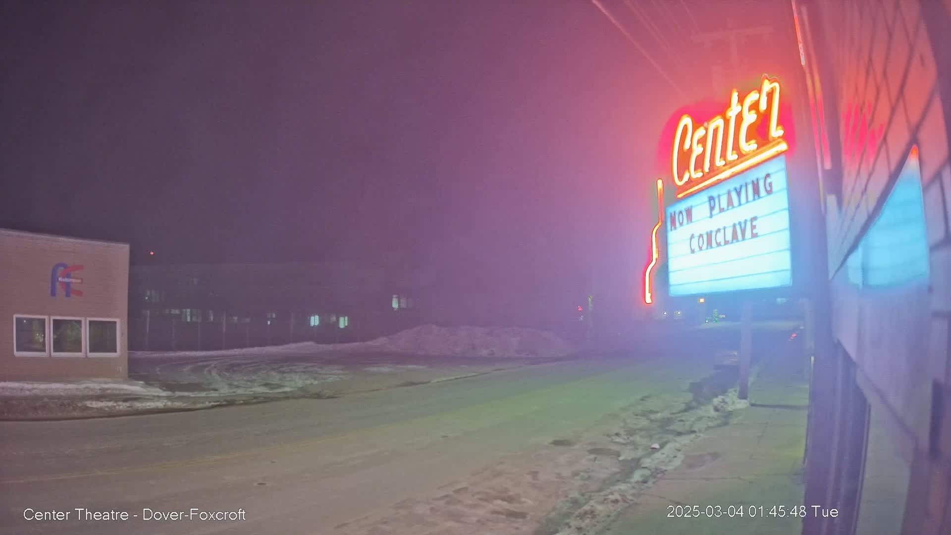 A snow-covered street at night is dimly lit by a neon-lit theater marquee advertising "Conclave."