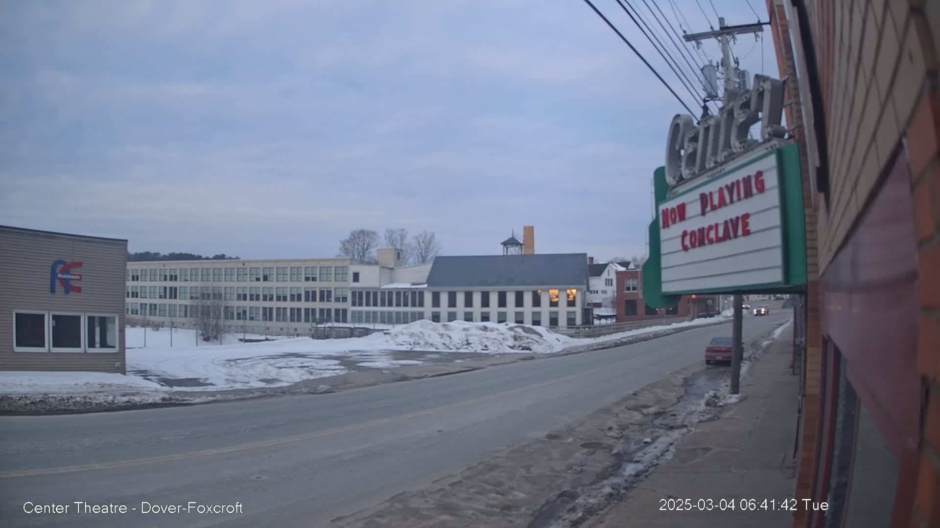 A mostly empty street scene on a cloudy day shows several buildings, including a large factory-like structure and a smaller building with a marquee, with patches of snow and ice on the ground and street.