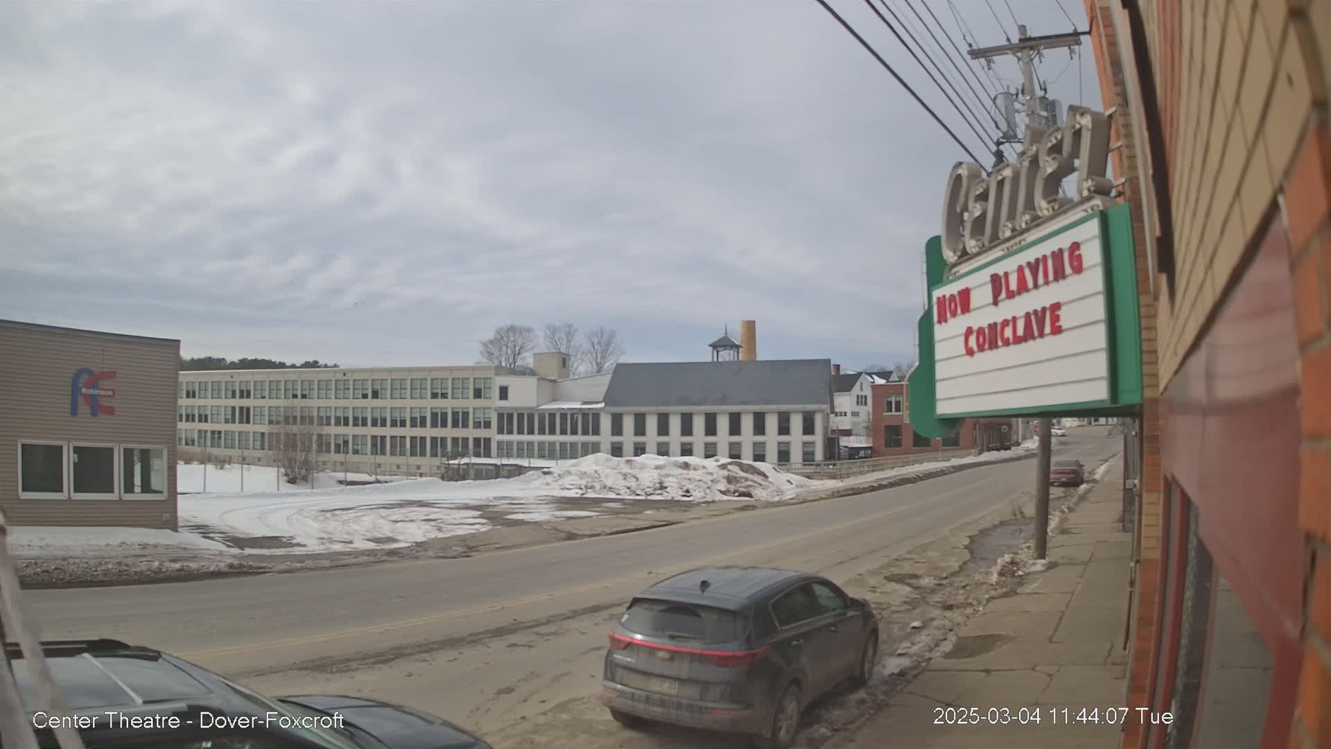 A partly cloudy day reveals a street scene with snow on the ground and parked cars, alongside multi-story buildings and a movie theater marquee.