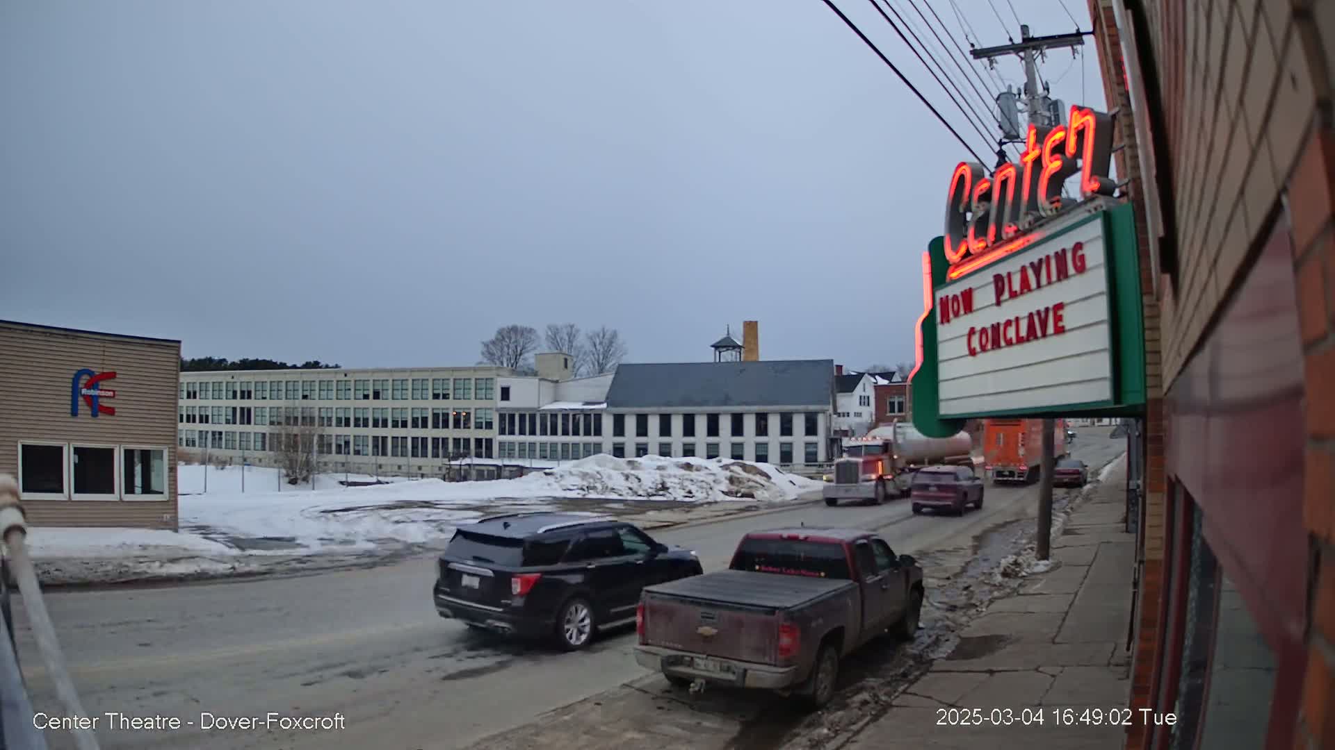 A street scene under an overcast sky shows several vehicles driving past a large building and a movie theater marquee advertising "Conclave."