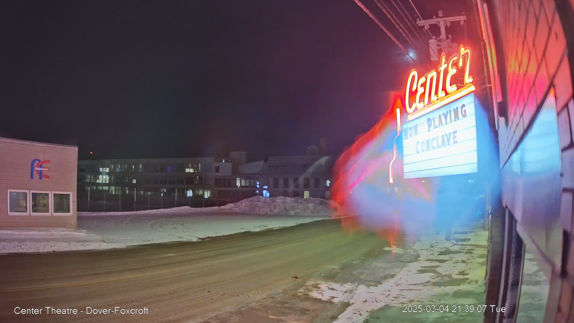 A snow-covered street at night with a movie theater marquee displaying "Now Playing Conclave" and illuminated buildings in the background.