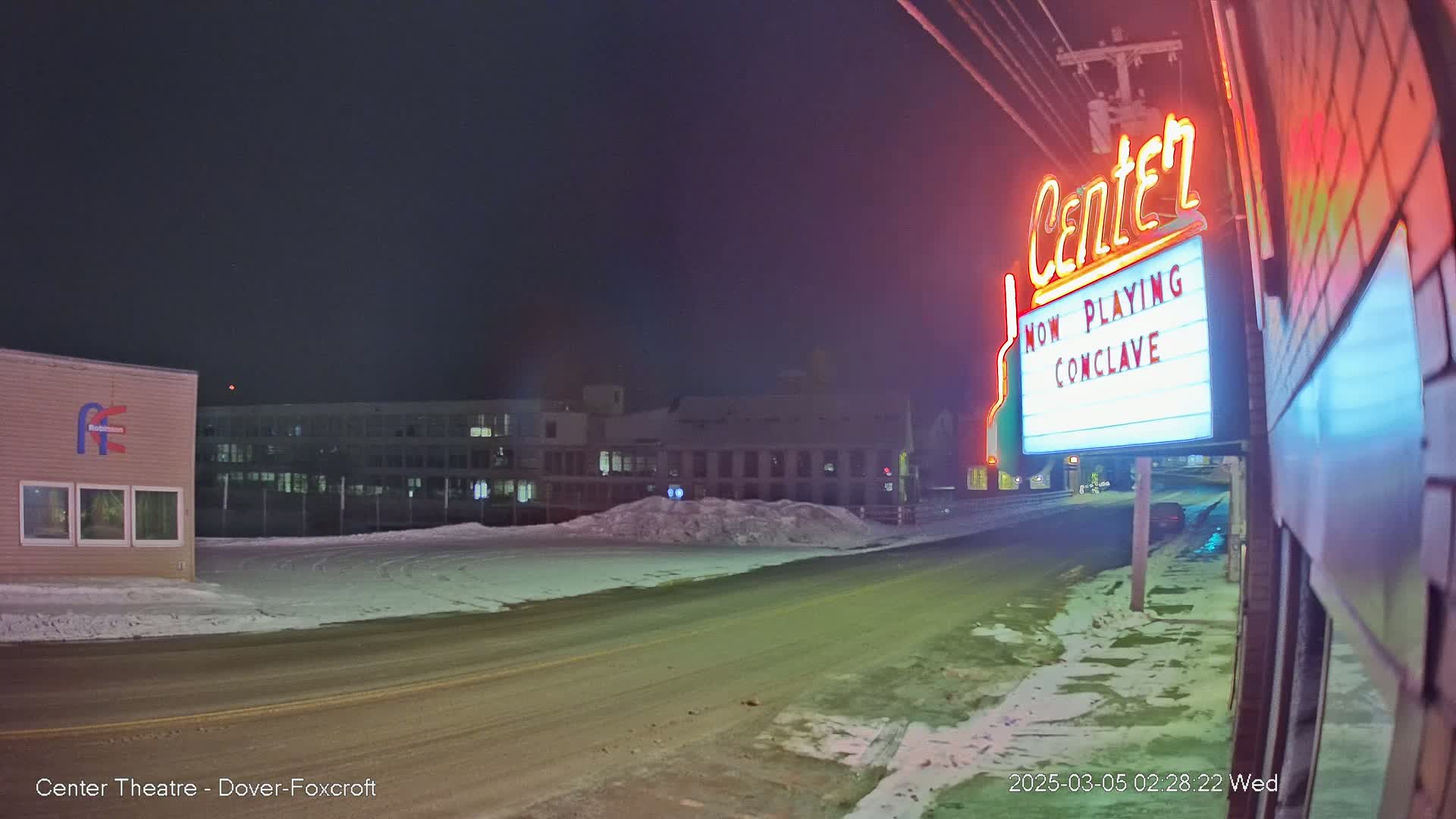 A snow-covered street at night is seen from a low angle, with a neon sign for a theater advertising a movie, and buildings in the background.