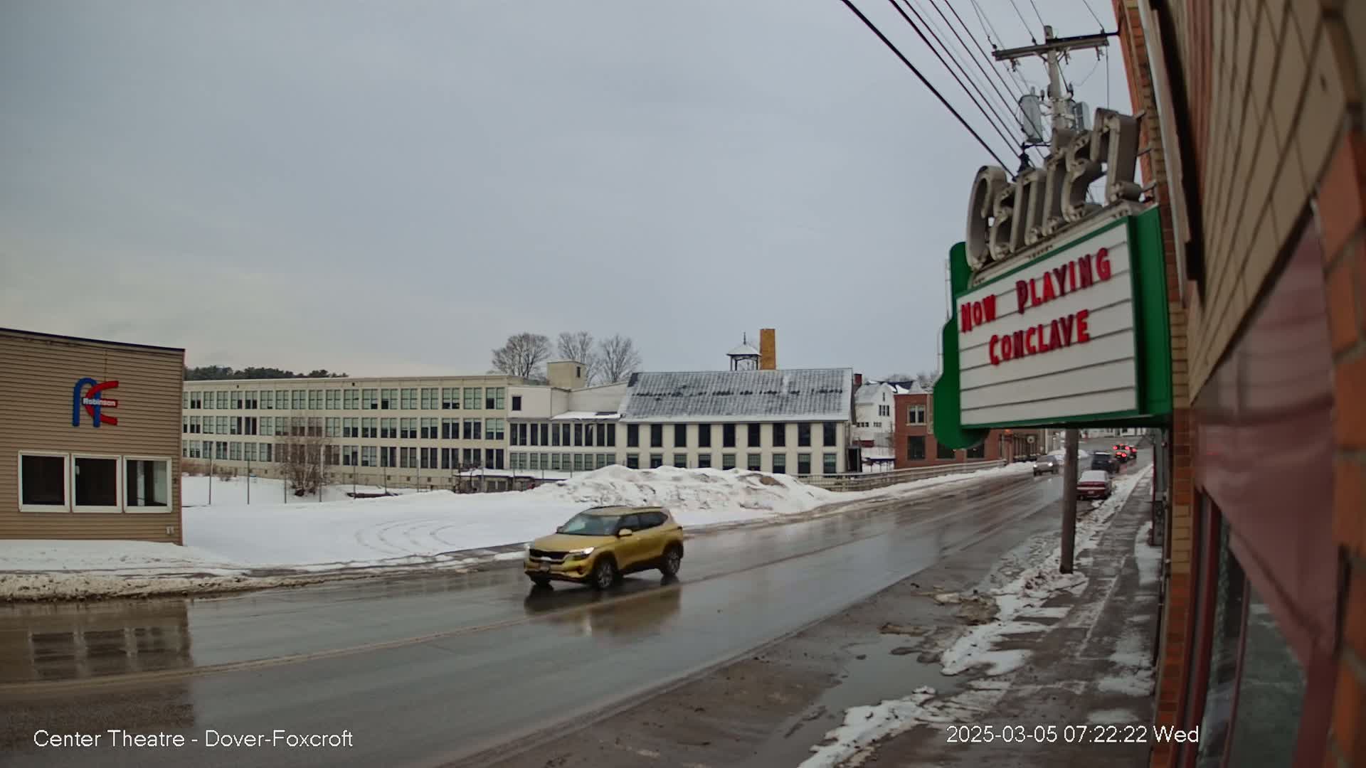 A gold SUV drives down a wet, snow-covered street past a multi-story building and a movie theater marquee under an overcast sky.