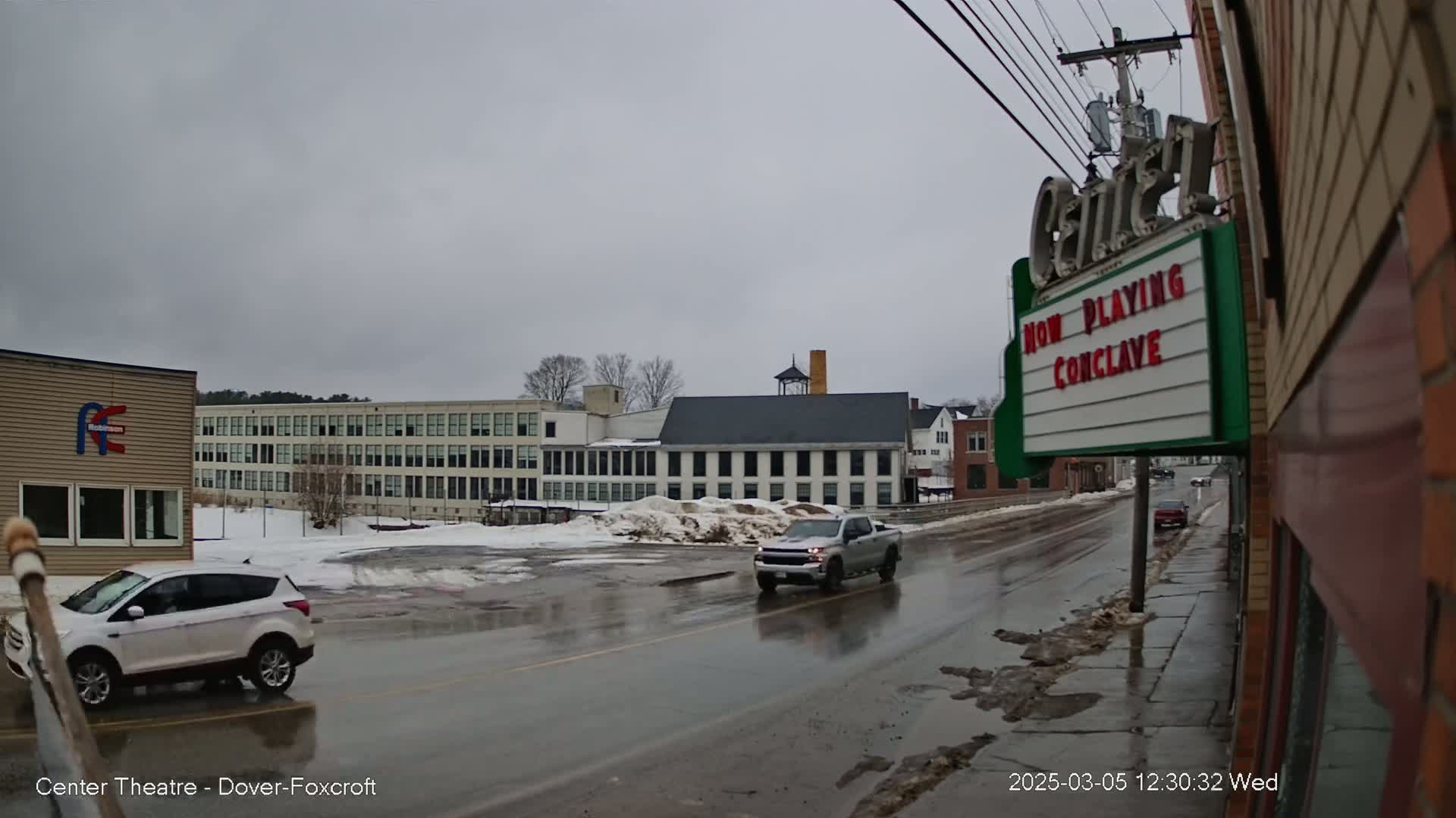 A wet street scene on an overcast day shows a pickup truck and SUV driving past a large, light-colored building and a movie theater marquee.