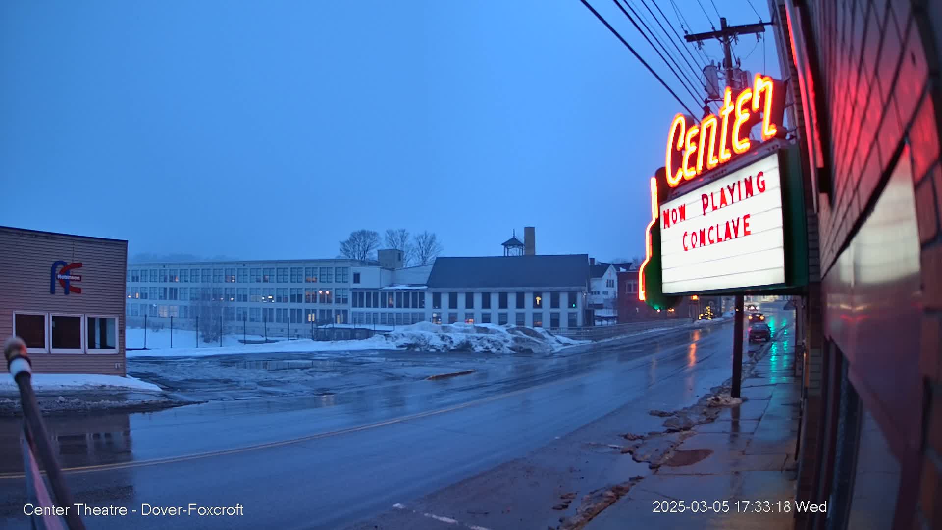A wet street scene at twilight shows a movie theater marquee advertising "Conclave," with buildings and snow-covered ground in the background.