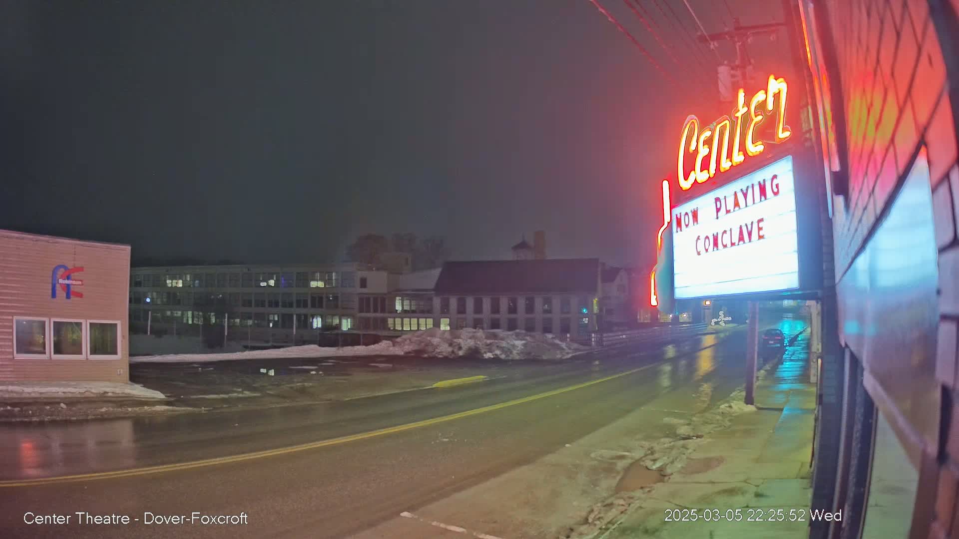 A wet, nighttime street scene shows a movie theater marquee advertising "Conclave," with snow banks and buildings in the background under a dark sky.