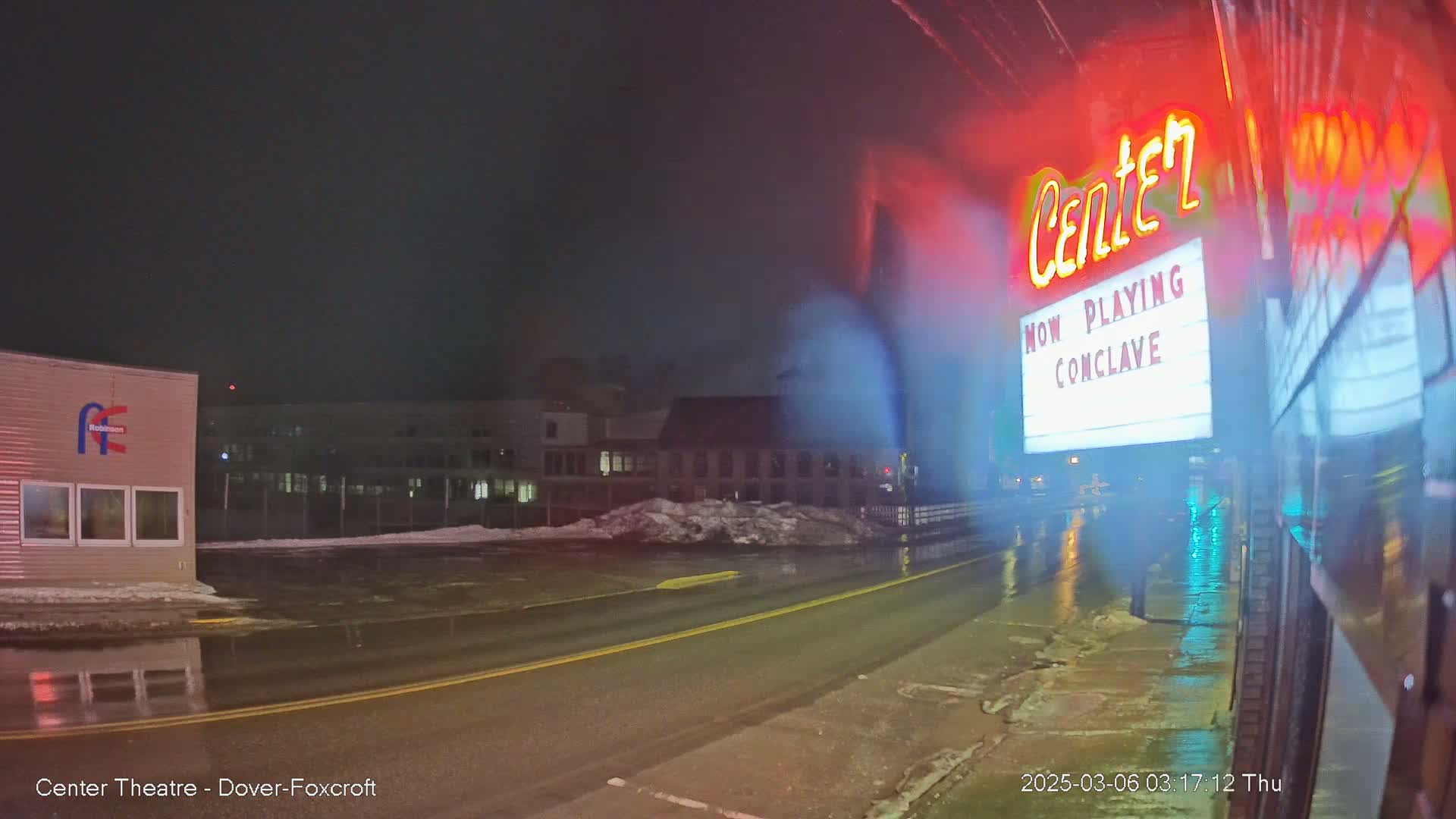A wet, nighttime street scene shows a movie theater marquee advertising "Conclave," with snow banks and buildings in the background.
