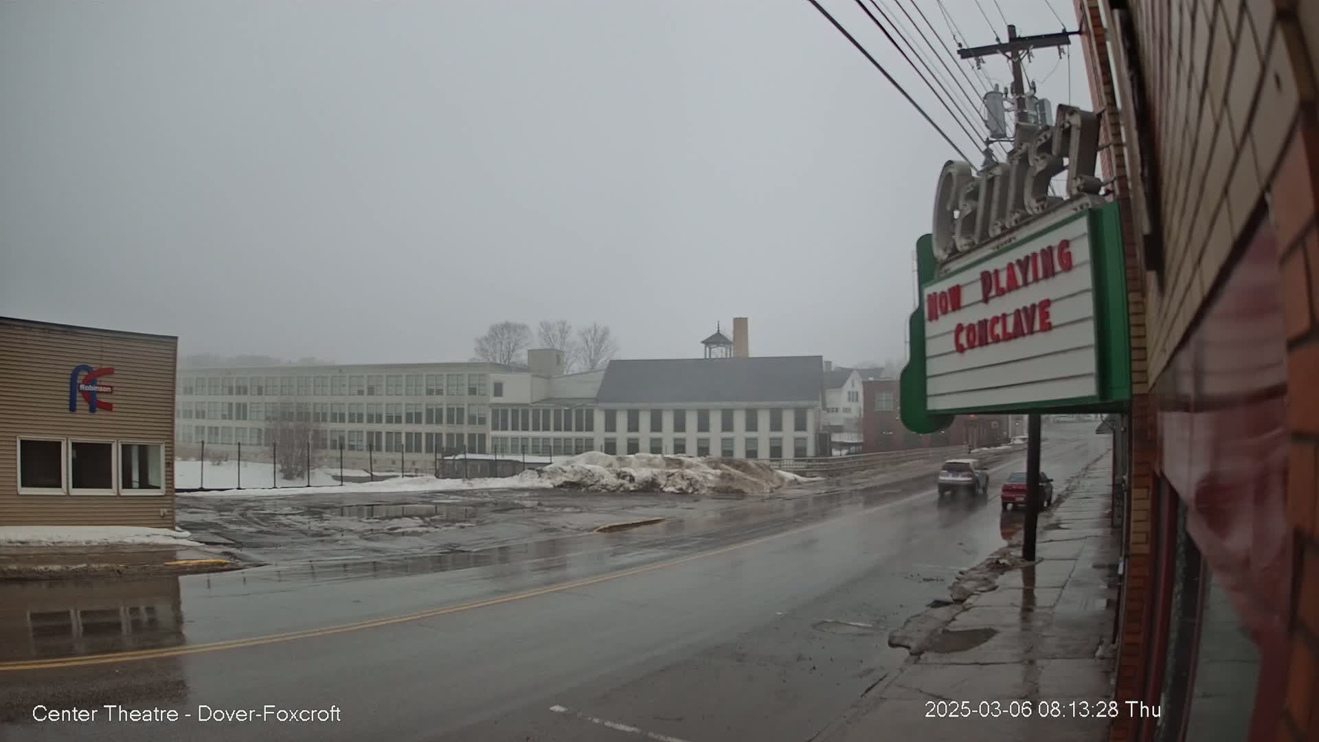 A wet, overcast street scene shows a multi-story building in the distance and a marquee advertising a movie, with a few cars driving on a wet road.