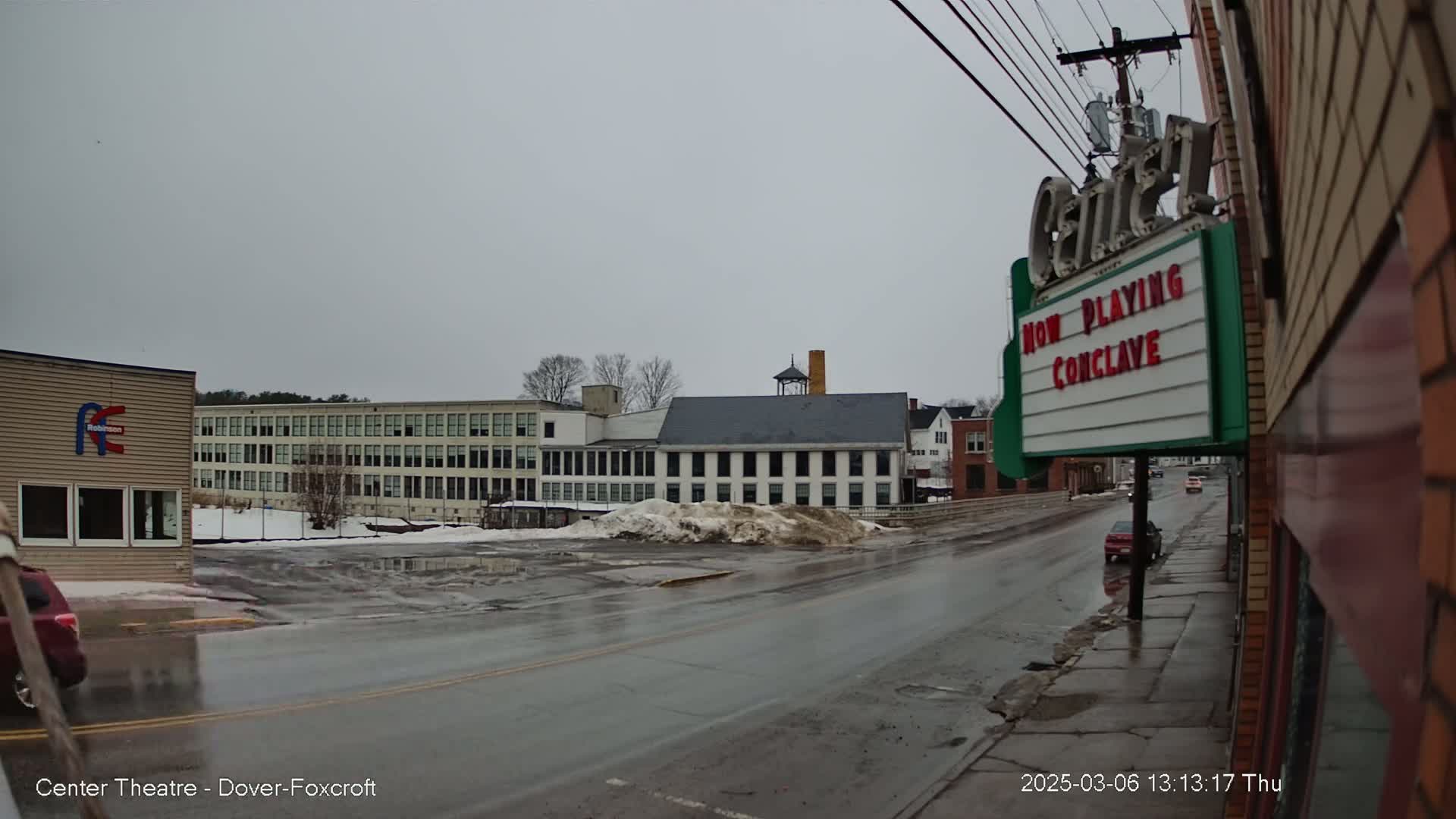 A wet street scene on an overcast day shows several buildings, including a large factory-like structure and a movie theater displaying a marquee.