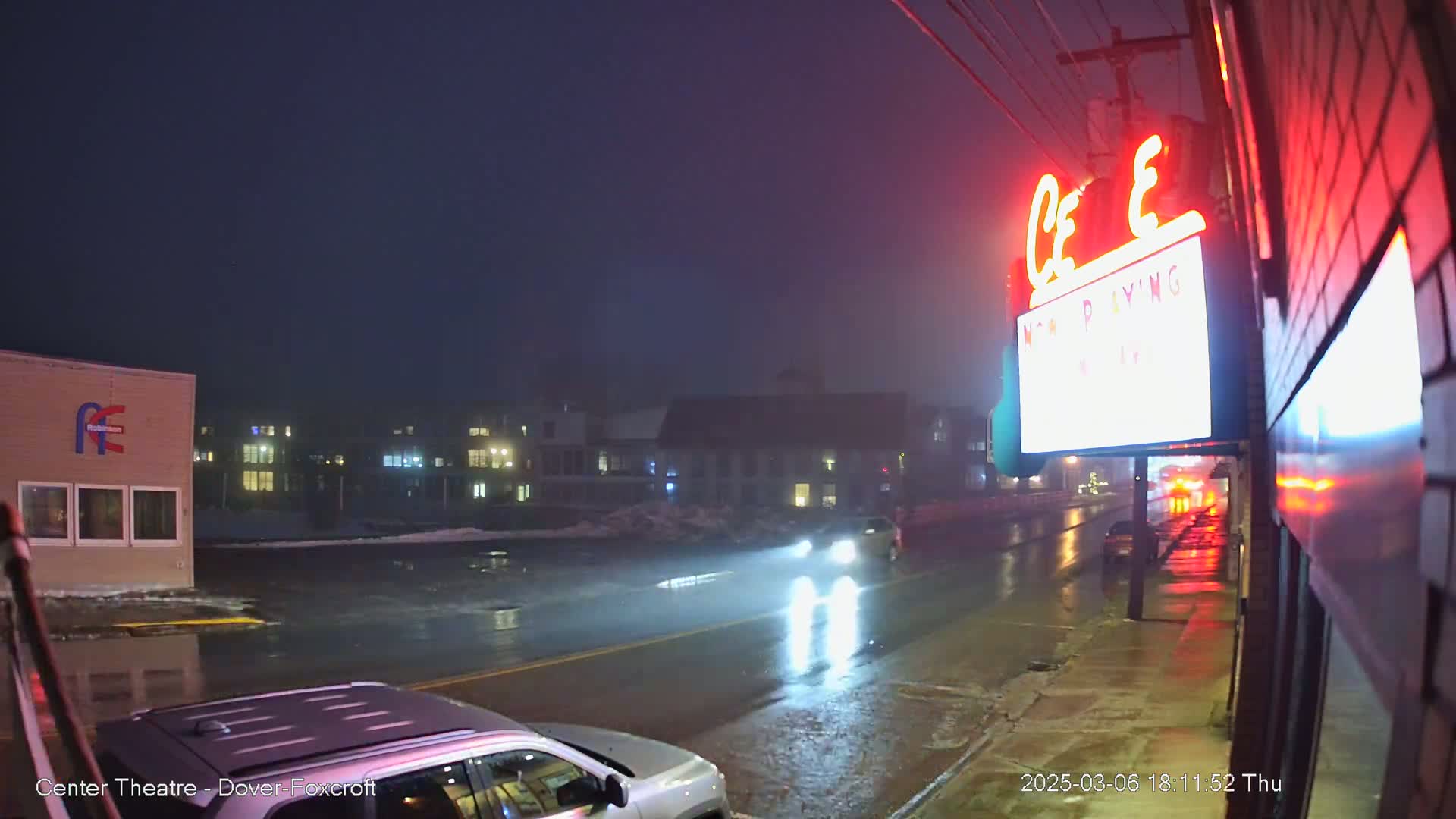 A wet street at night, illuminated by headlights and neon signs, shows a person walking near a movie theater.