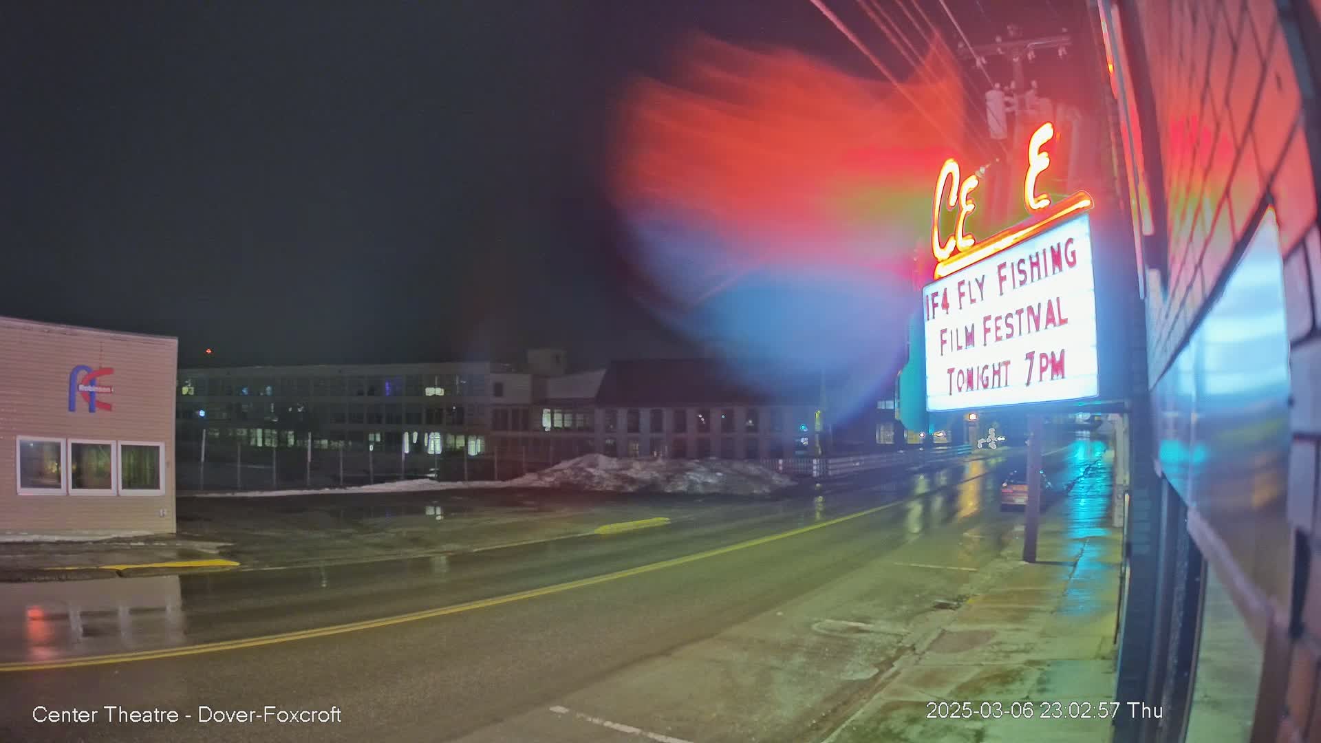 A wet, nighttime street scene shows a marquee advertising a film festival, with buildings and snow visible in the background under a dark sky.