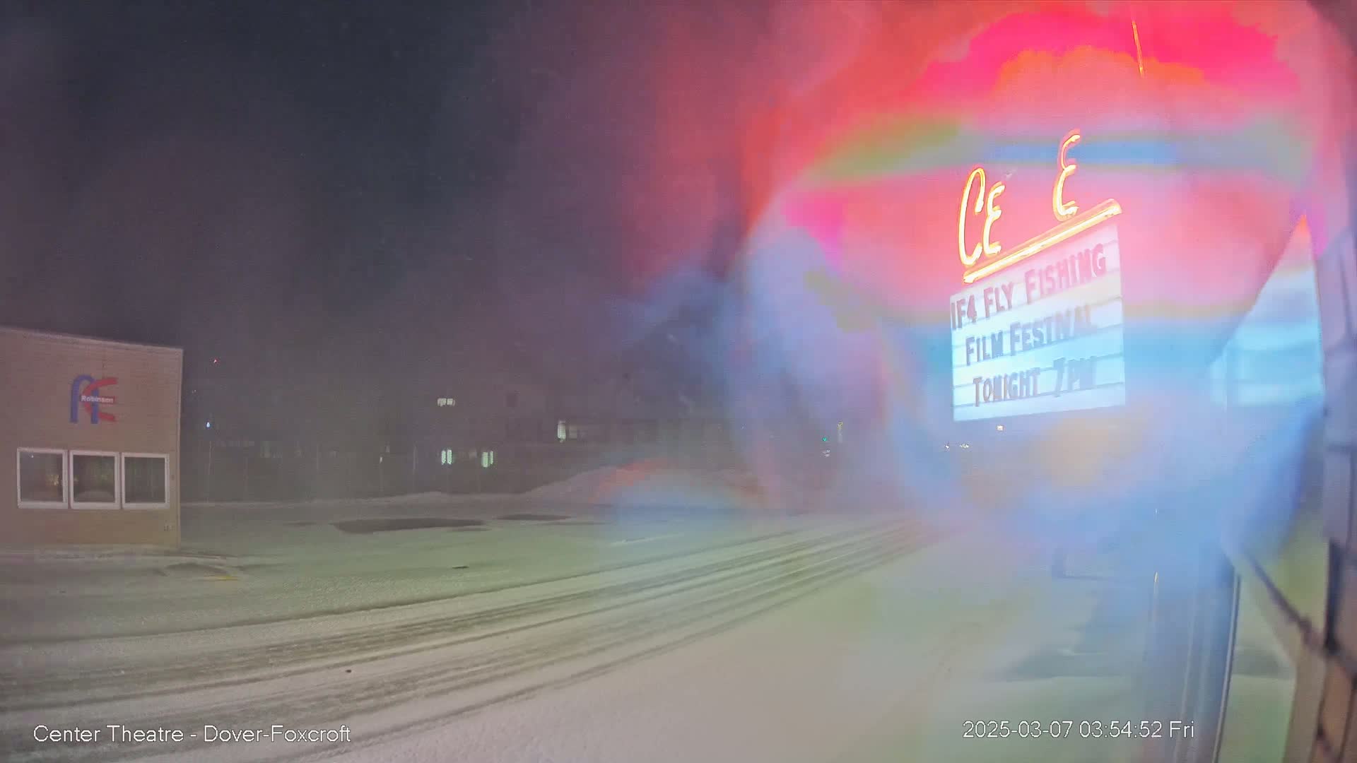 A snow-covered parking lot and buildings are visible through a hazy, snow-filled atmosphere at night, with a brightly lit marquee in the background.