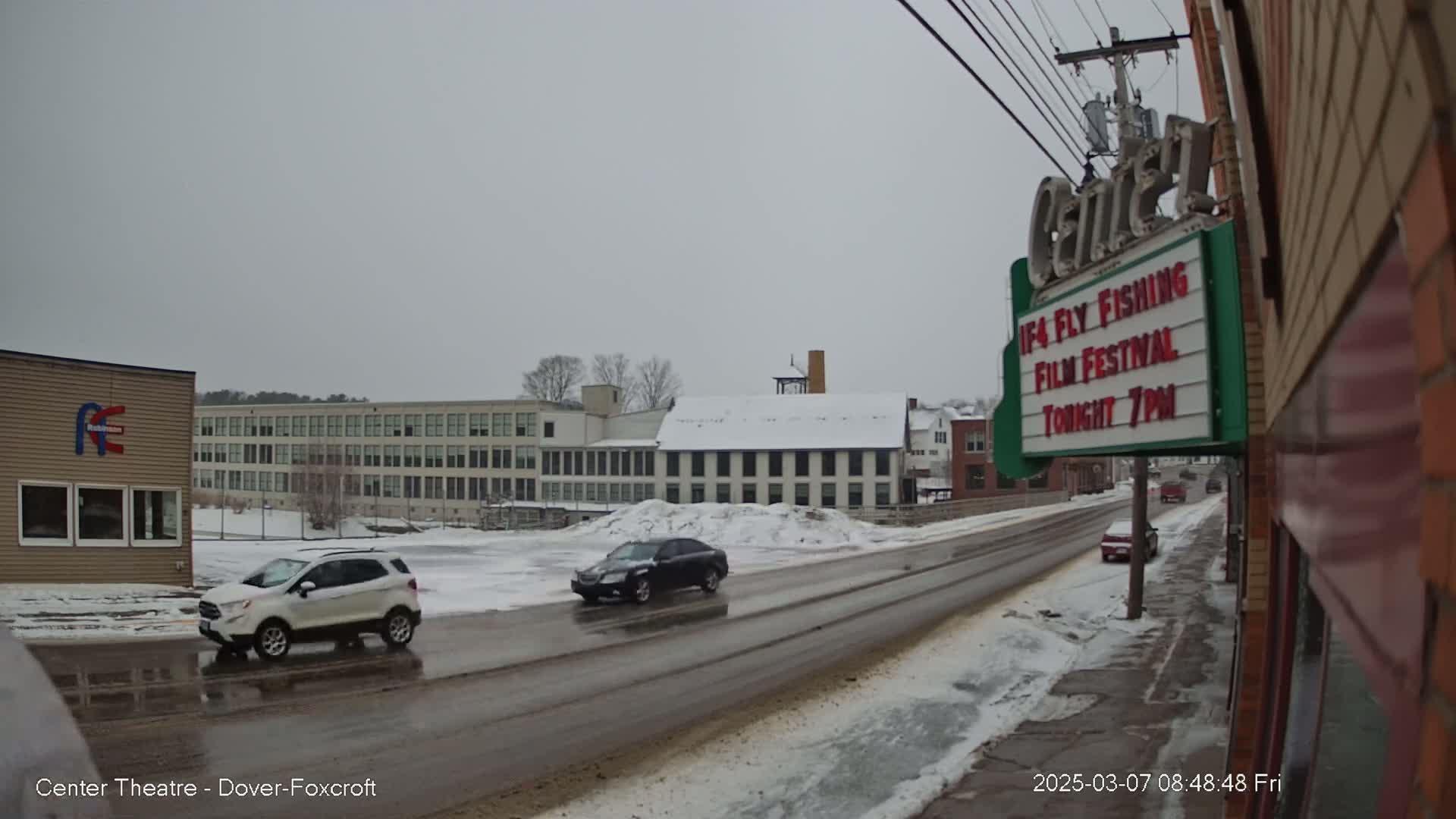 A snow-covered street scene shows cars driving past a multi-story building and a theater marquee advertising a film festival.