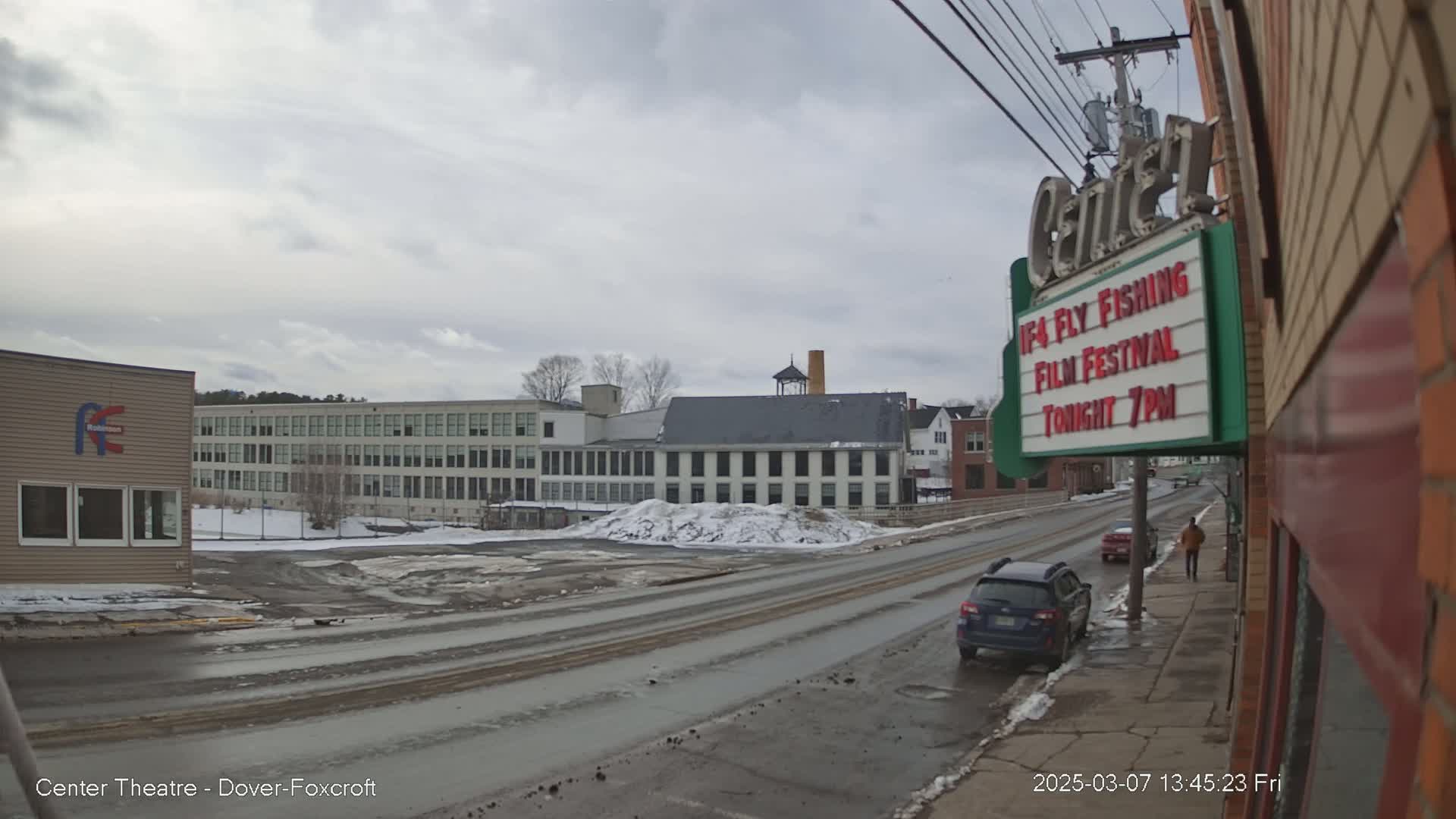 A mostly cloudy day reveals a street scene with snow on the ground and buildings, including a large multi-story structure and a smaller brick building with a marquee.