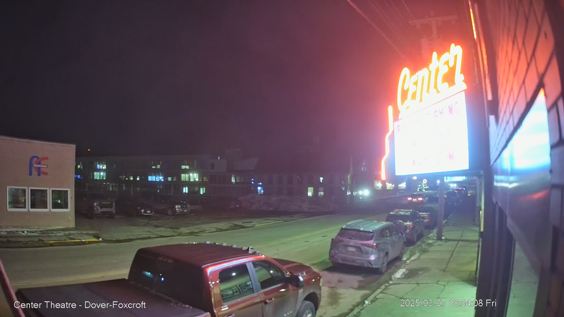 A nighttime street scene shows several parked cars and a red pickup truck near a building with a lit "Center" sign.