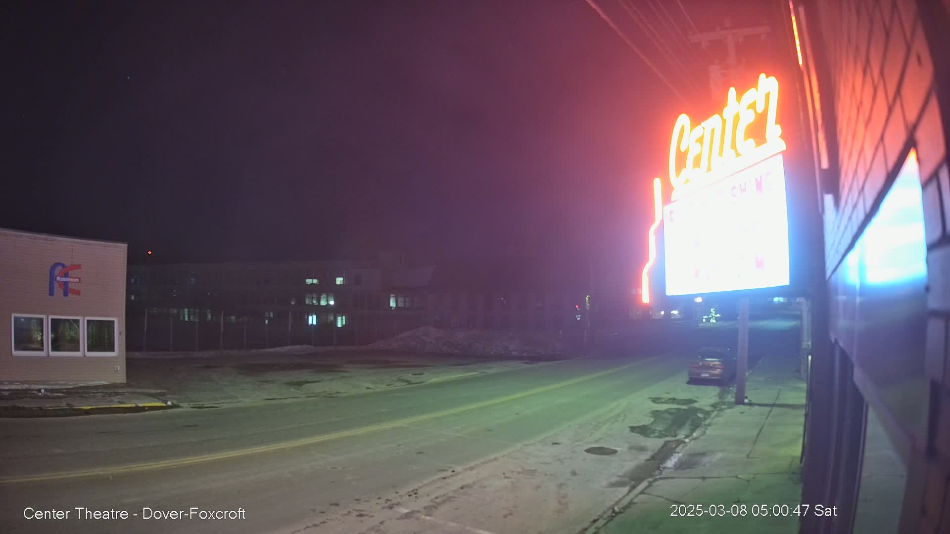 A mostly empty street at night, with snow on the ground, is seen under a dark sky; a neon sign for Center Theatre is visible on the right.