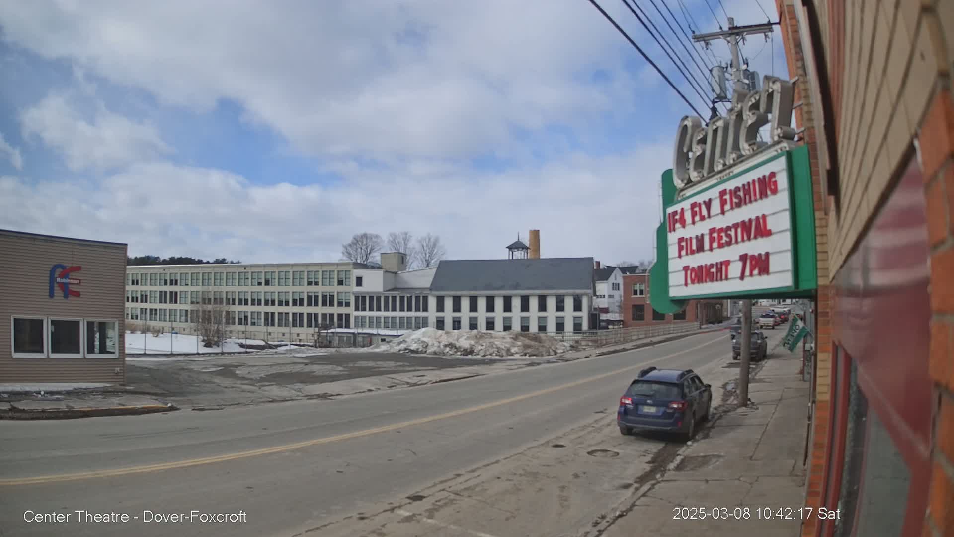 A mostly sunny day reveals a street scene with several buildings, including a large factory-style structure and a theater marquee advertising a film festival, with a few cars parked along the street.
